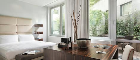 A modern hotel room with a large bed, a wooden desk, a chair, and a view of greenery outside.