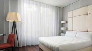 A modern hotel room with a large bed, a desk with a chair, a floor lamp, and a window with sheer curtains. - Sina The Gray (Milan)