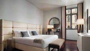 Ett hotellrum med en stor säng, sängbord, ett skrivbord, en stol, en spegel och utsikt över byggnader genom ett fönster.