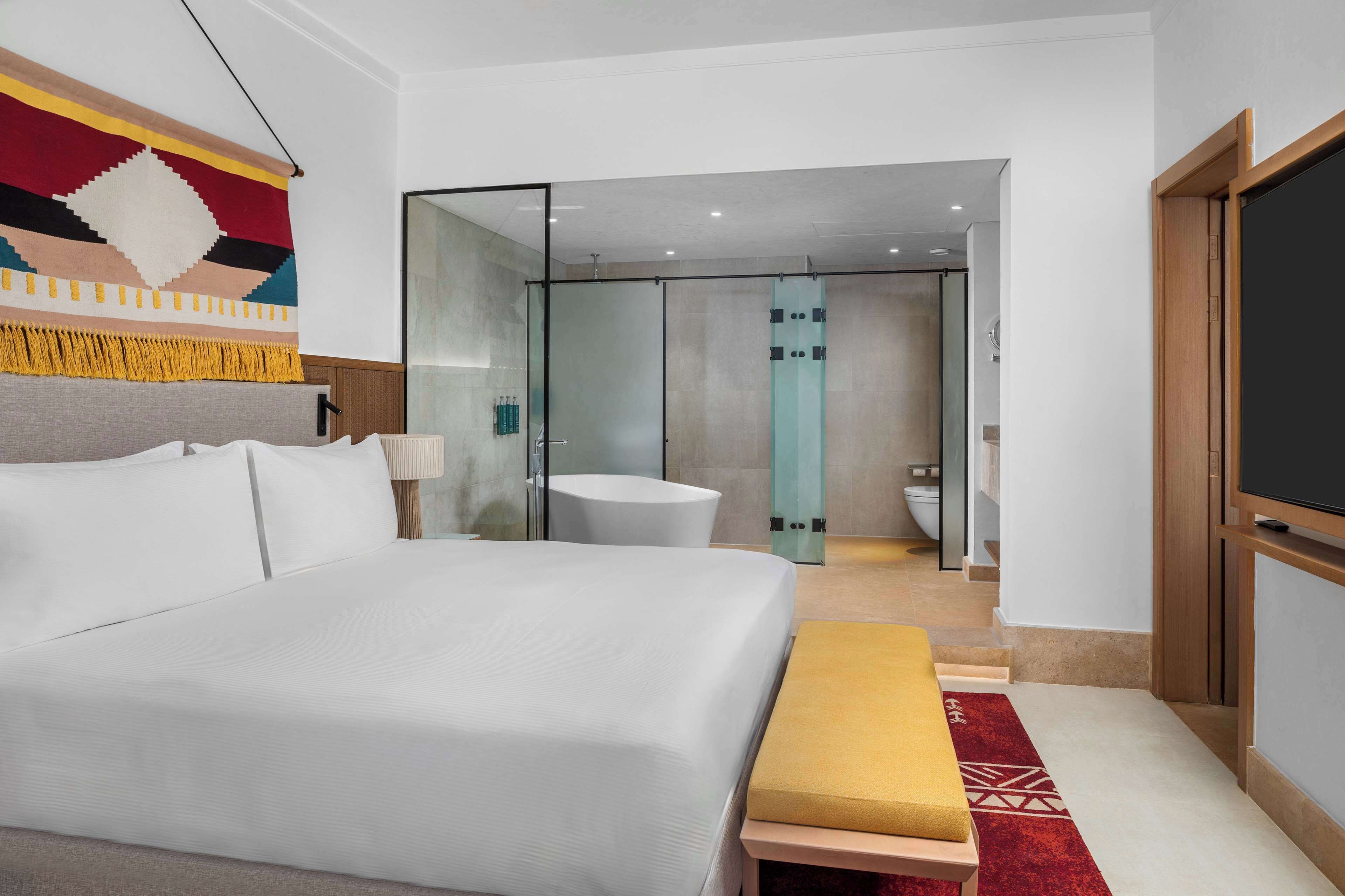 A modern hotel room with a large bed, a bench, a TV, and a bathroom visible through a glass door.