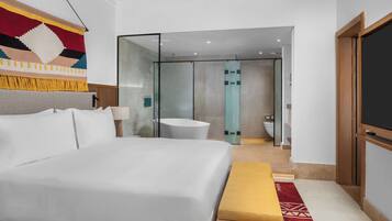Una habitación de hotel moderna con una cama grande, un banco, un televisor y un baño visible a través de una puerta de vidrio.