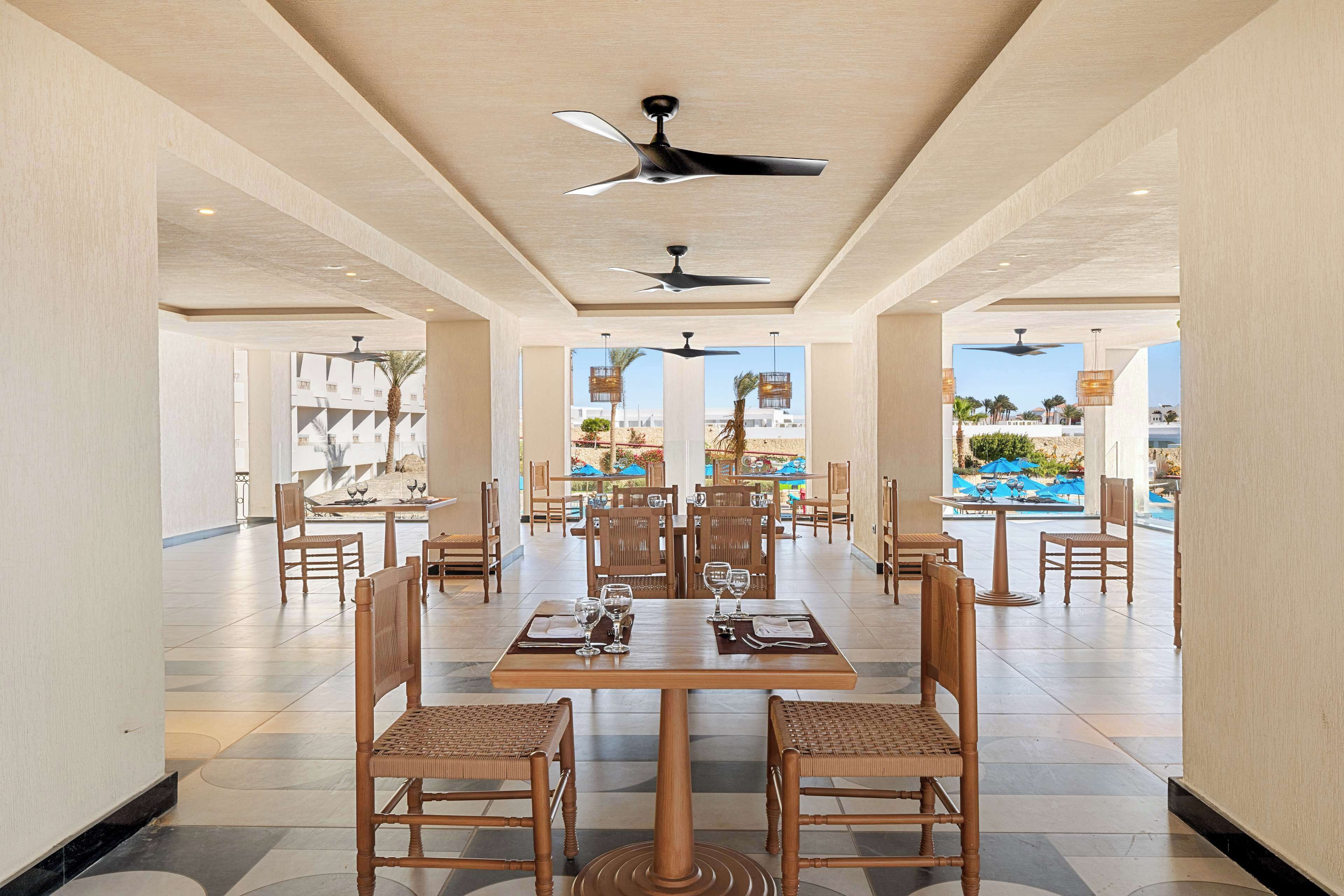 A spacious indoor dining area with multiple tables and chairs, ceiling fans, and large windows offering views of an outdoor pool area.