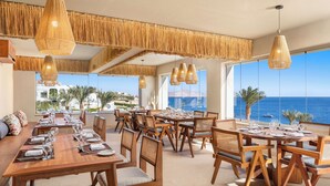 Un restaurante con mesas y sillas de madera, ventanas amplias con vista al mar y lámparas colgantes de mimbre.