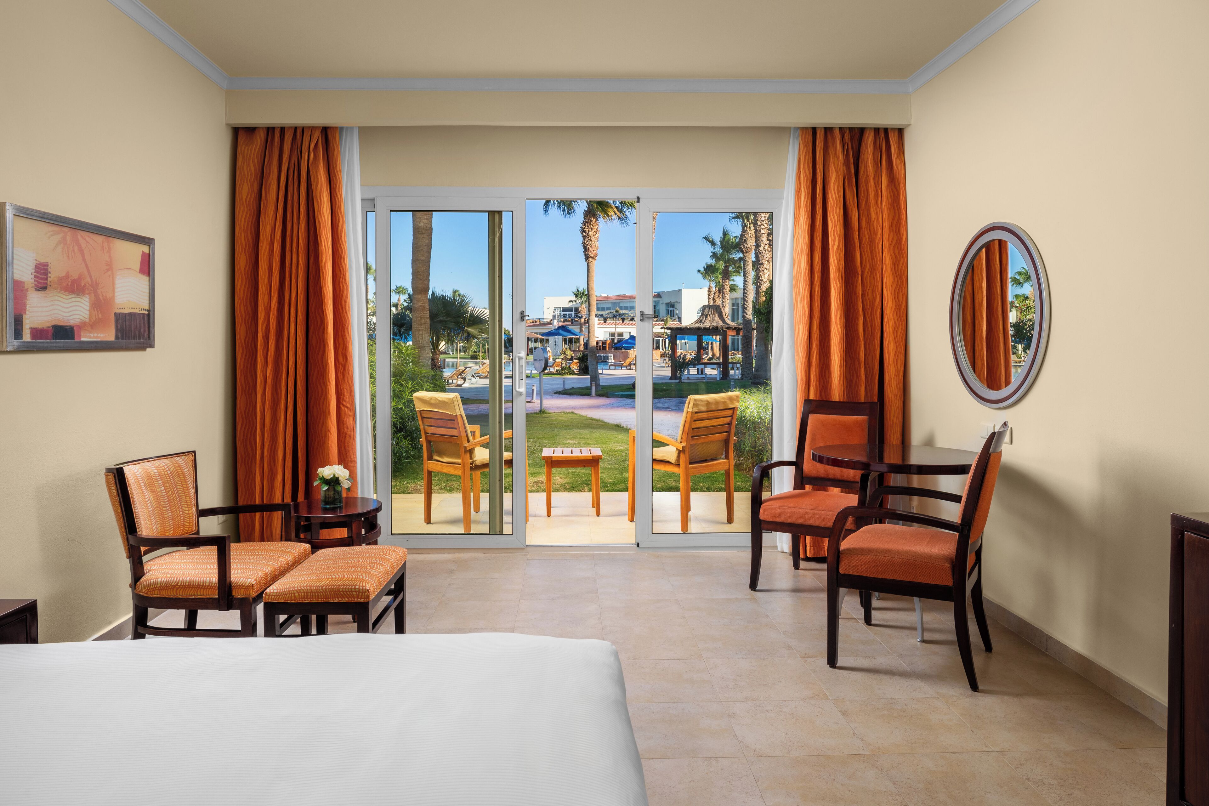 A hotel room with a bed, two chairs, a small table, and a view of a pool and palm trees through large glass doors.
