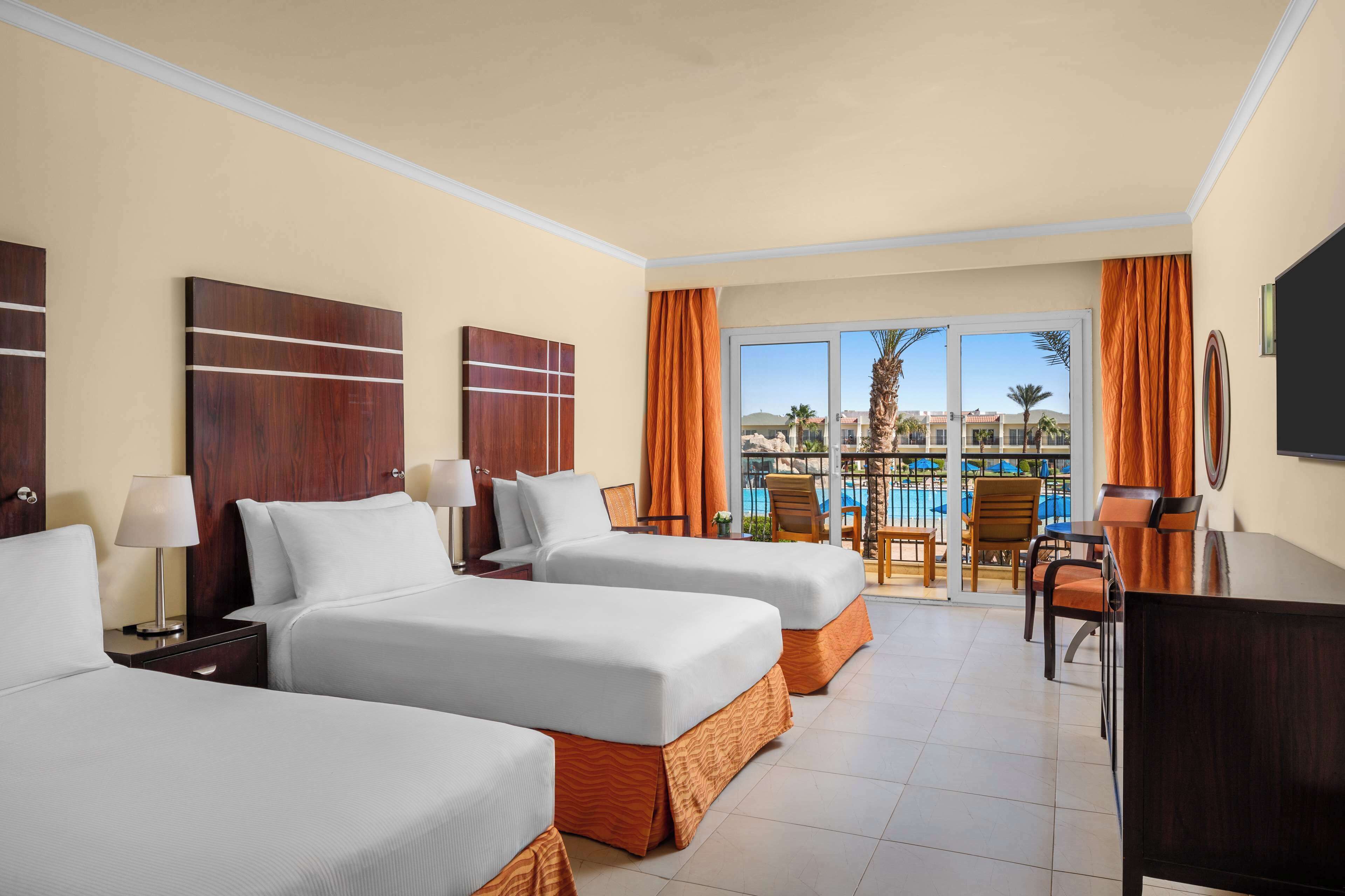Habitación de hotel con dos camas, un escritorio y vistas a una piscina y palmeras.