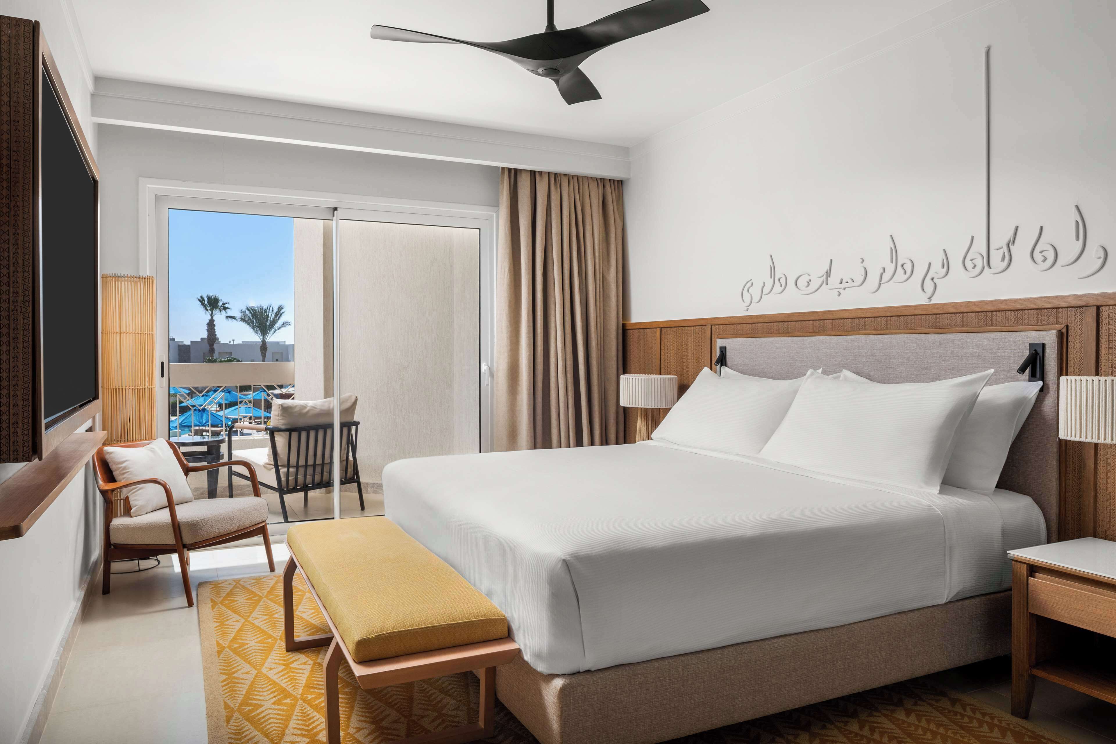 A hotel room with a bed, a desk, a chair, a window with a view of palm trees, and a ceiling fan.