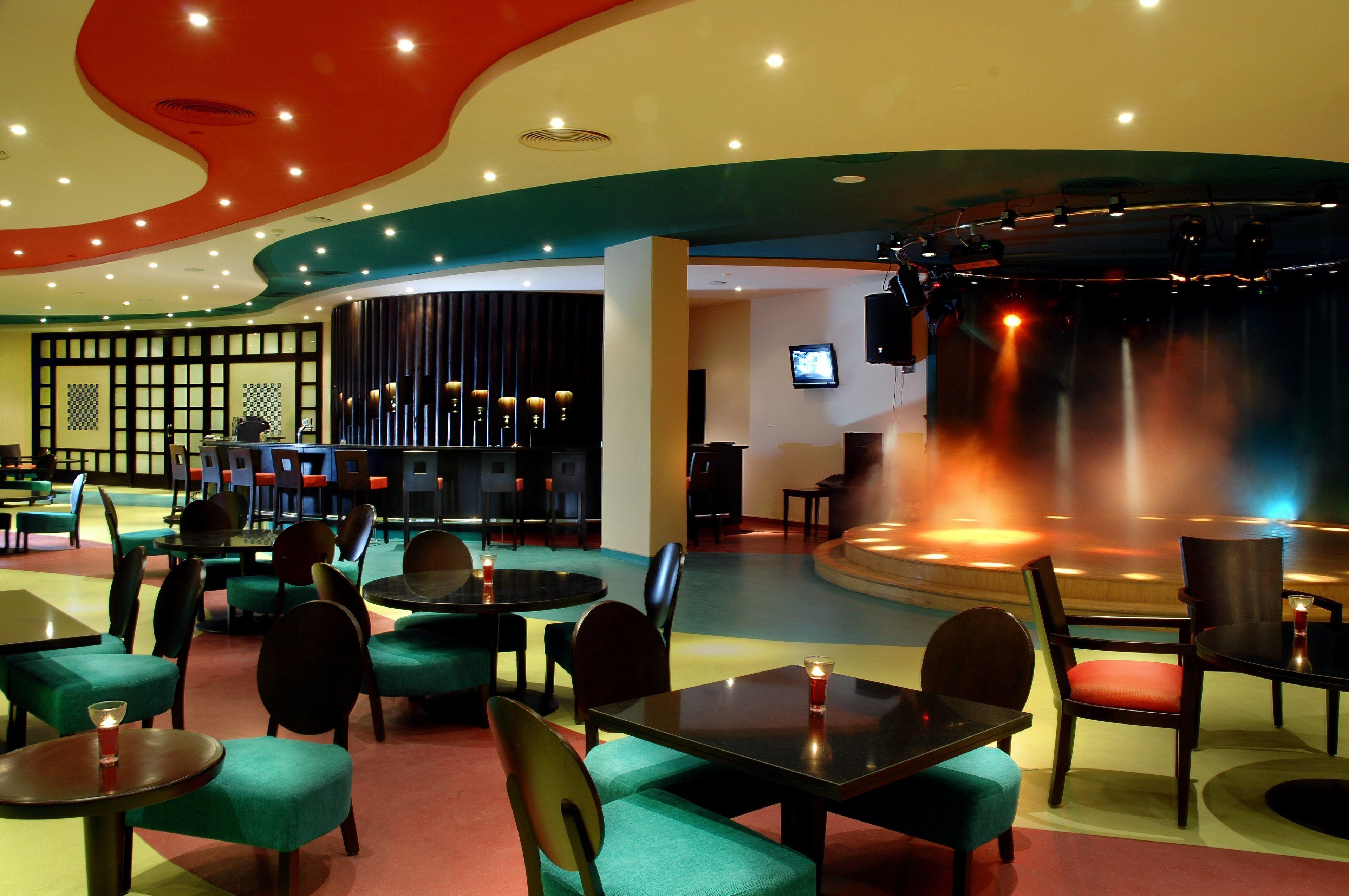 A restaurant with round tables and chairs, a stage with a screen, and a bar area with stools.