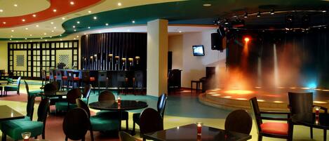 A restaurant with round tables and chairs, a stage with a screen, and a bar area with stools.