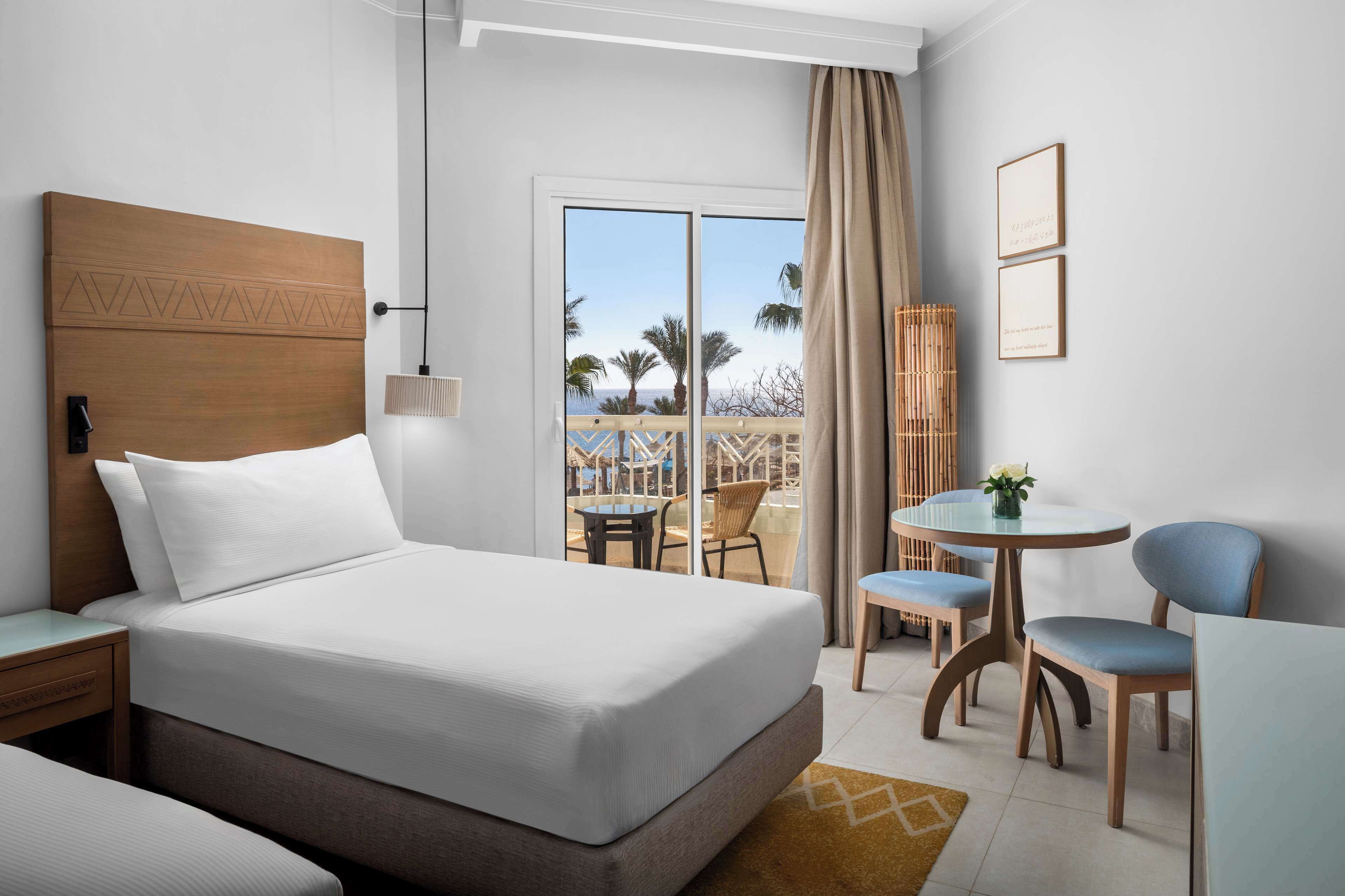 Ein Hotelzimmer mit einem großen Bett, einem kleinen Tisch mit Stühlen, einem Balkon mit Blick auf Palmen und einem Holz-Kopfstück.