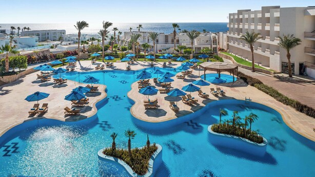 Zona de piscina en un resort, con varios lugares para tomar el sol y sombrillas, rodeada de palmeras y edificios.