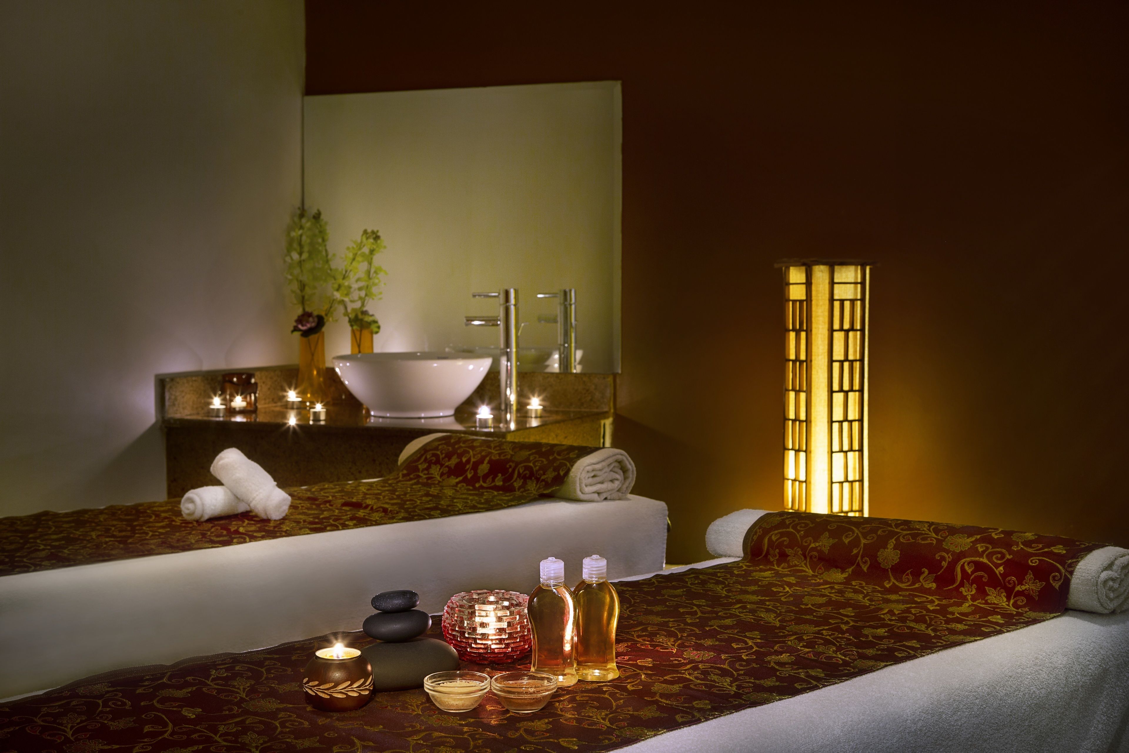 A well-lit spa room with a large mirror, a sink, and a bed with a red patterned bedspread.