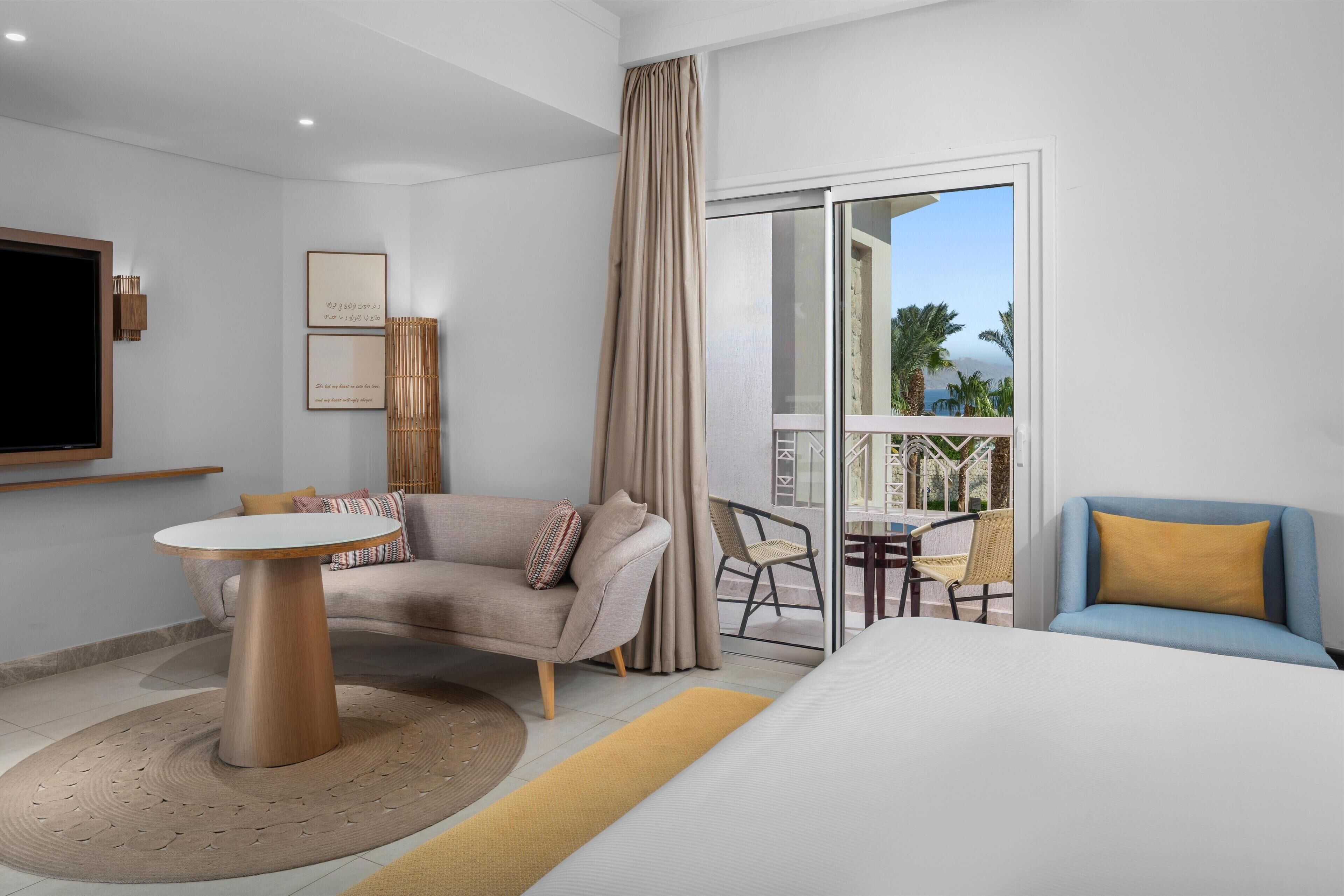 Une chambre d’hôtel moderne avec un grand lit, un canapé, une petite table ronde et un balcon offrant une vue sur les palmiers.