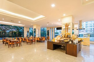 A spacious hotel dining area with multiple tables and chairs, a buffet counter, and large windows offering views of the cityscape.