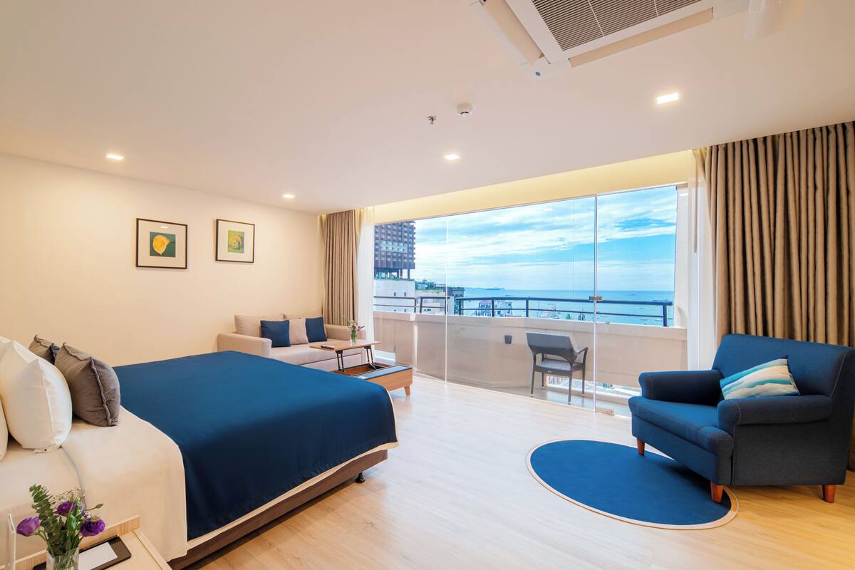 A modern hotel room with a large bed, a sofa, a chair, and a balcony with a view of the city and the sea.