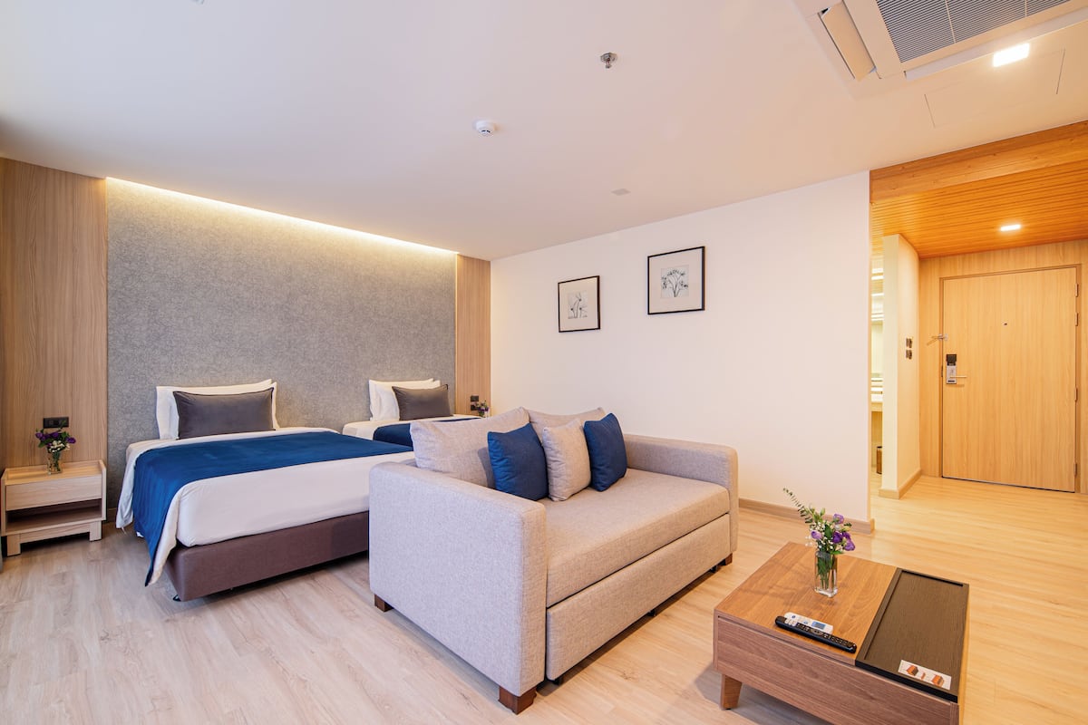 A modern hotel room with a bed, a sofa, a bedside table, and a wooden door.