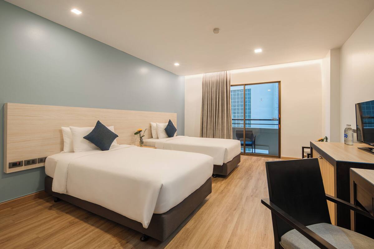 A hotel room with two beds, a desk, a chair, and a view of a building through a window.
