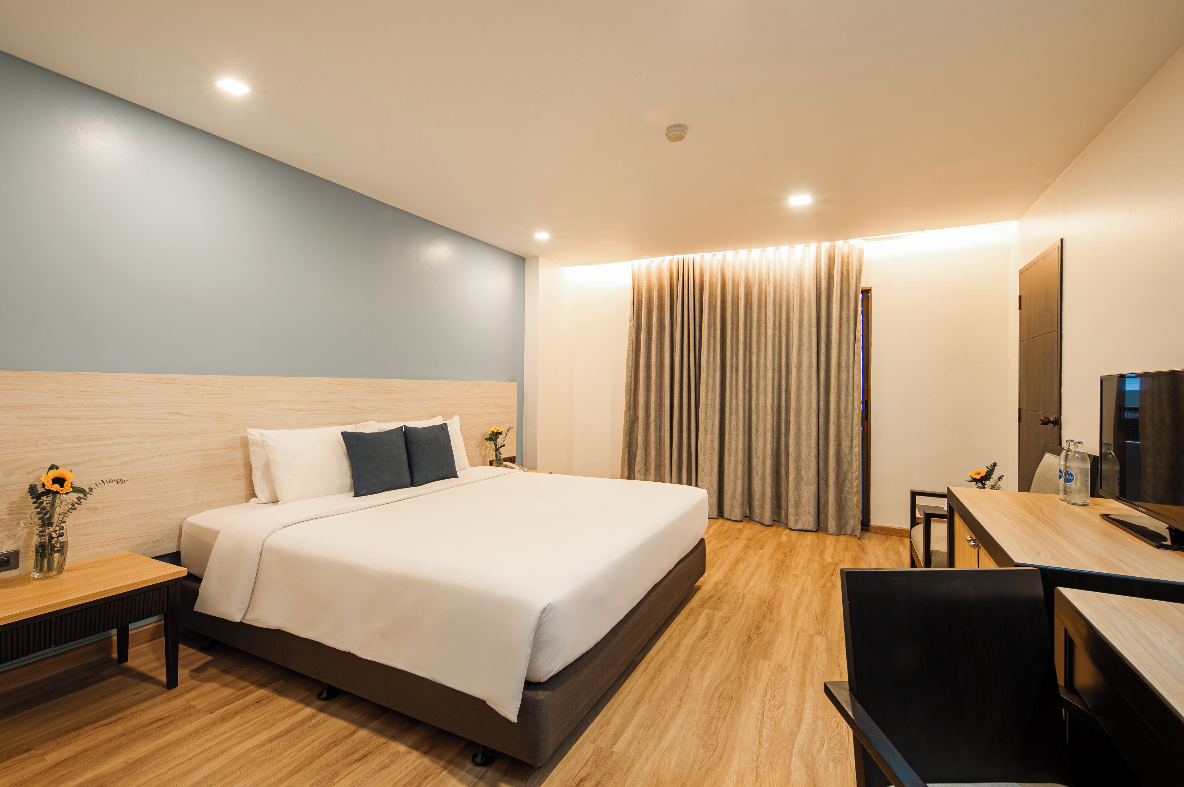 Ein modernes Hotelzimmer mit einem großen Bett, einem Holztisch, einem Sessel, einem Fernseher und einem Fenster mit Vorhängen.