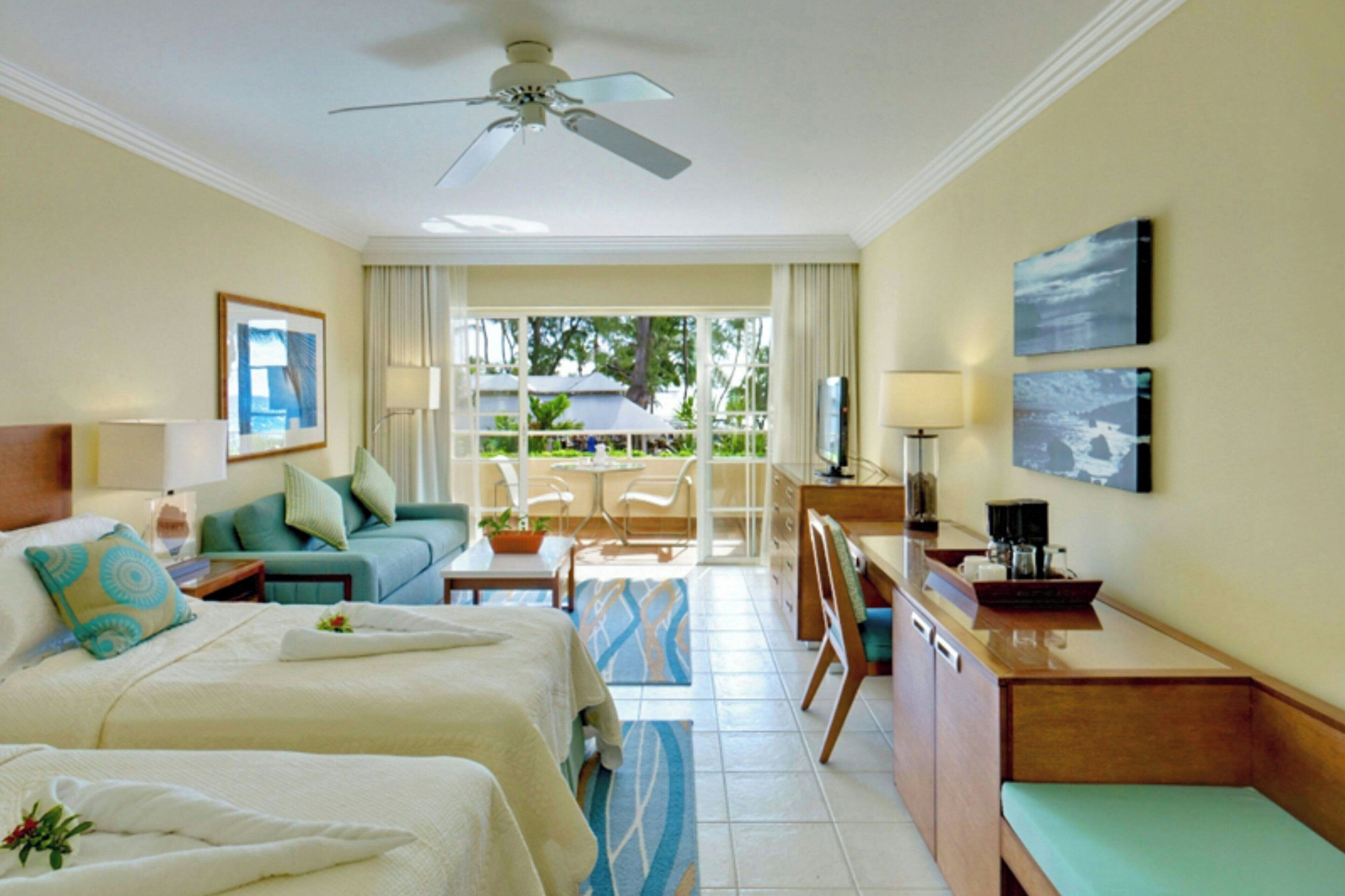 Habitación de hotel con dos camas, un escritorio, una silla, un sofá, un balcón con vistas y un ventilador de techo.