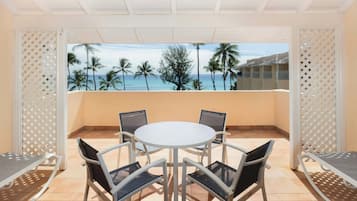 A balcony with a white table and chairs, overlooking a beach with palm trees and a clear sky.