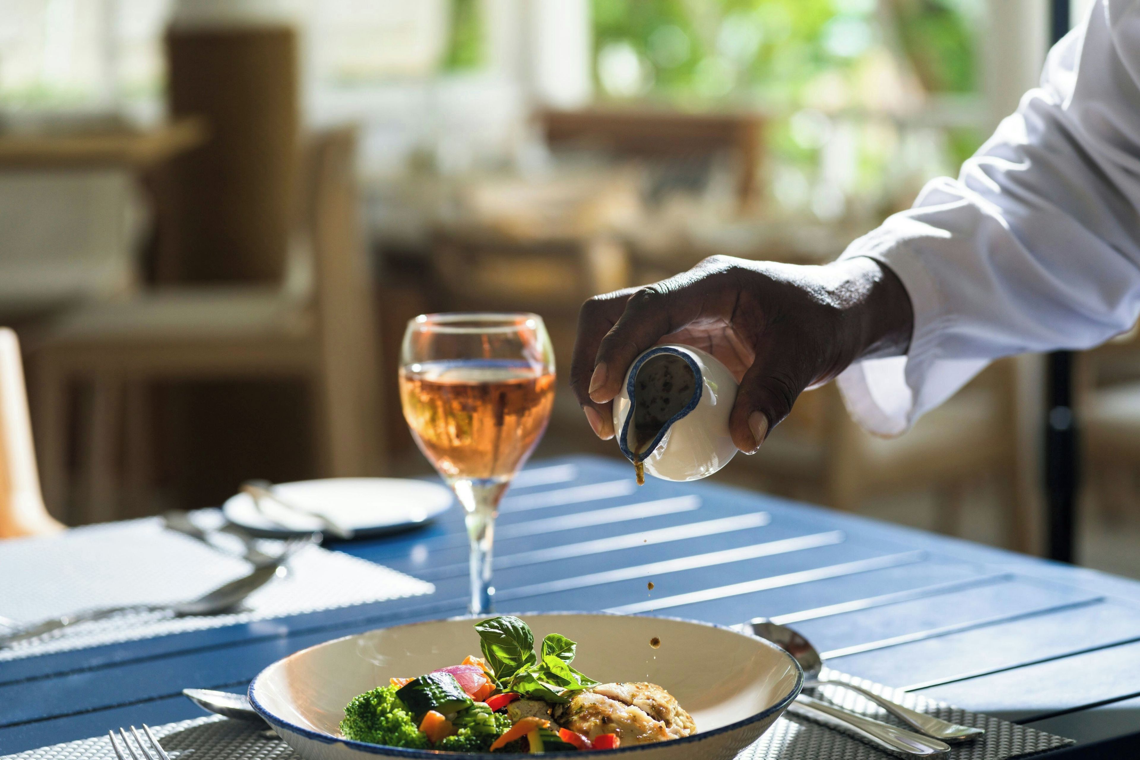 A dining table with a plate of food, a glass of wine, and a hand pouring salt.