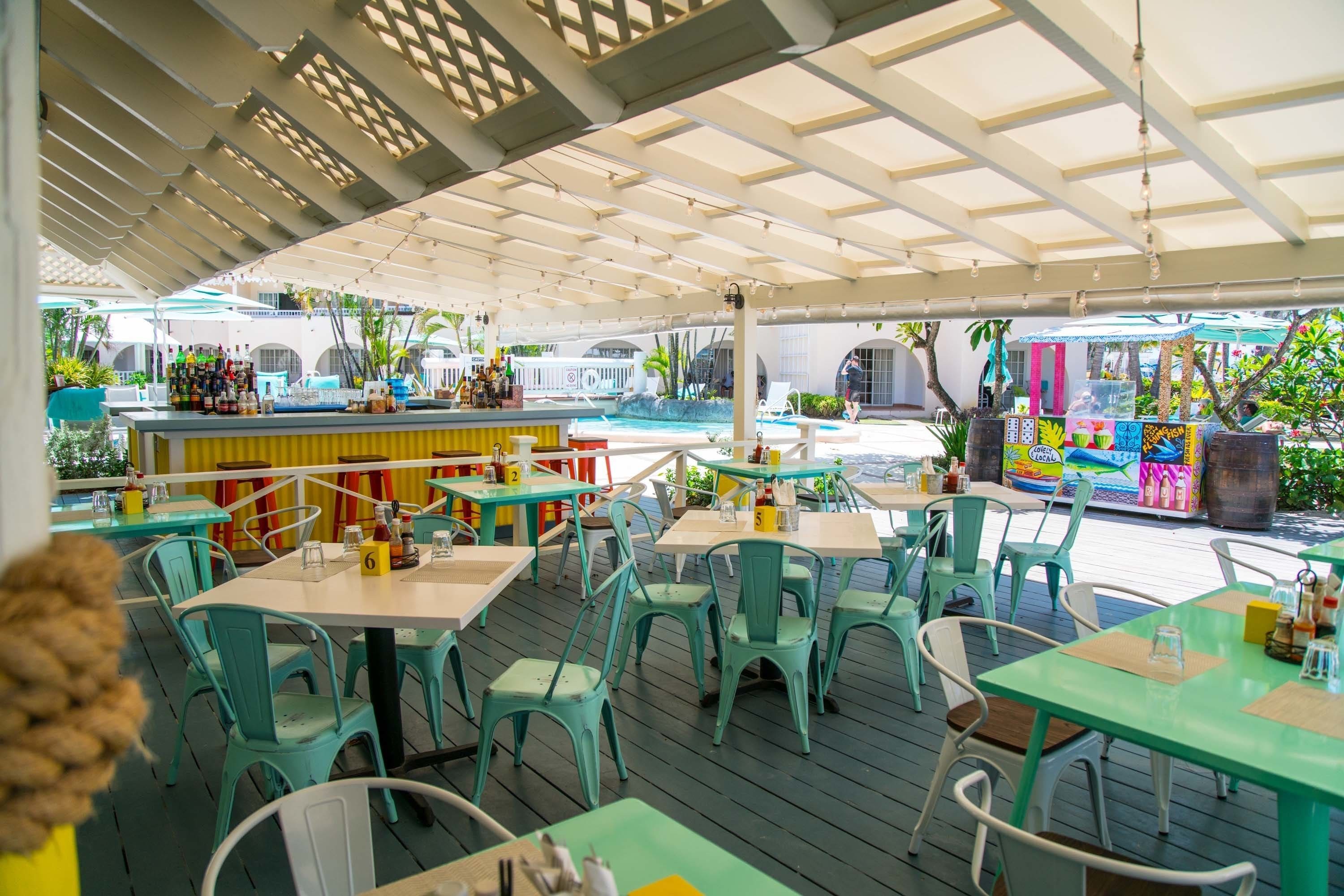 Een terras met turquoise tafels en stoelen, een bar met een kleurrijke muurschildering en een zwembad op de achtergrond.