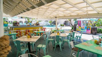 Een terras met turquoise tafels en stoelen, een bar met een kleurrijke muurschildering en een zwembad op de achtergrond.