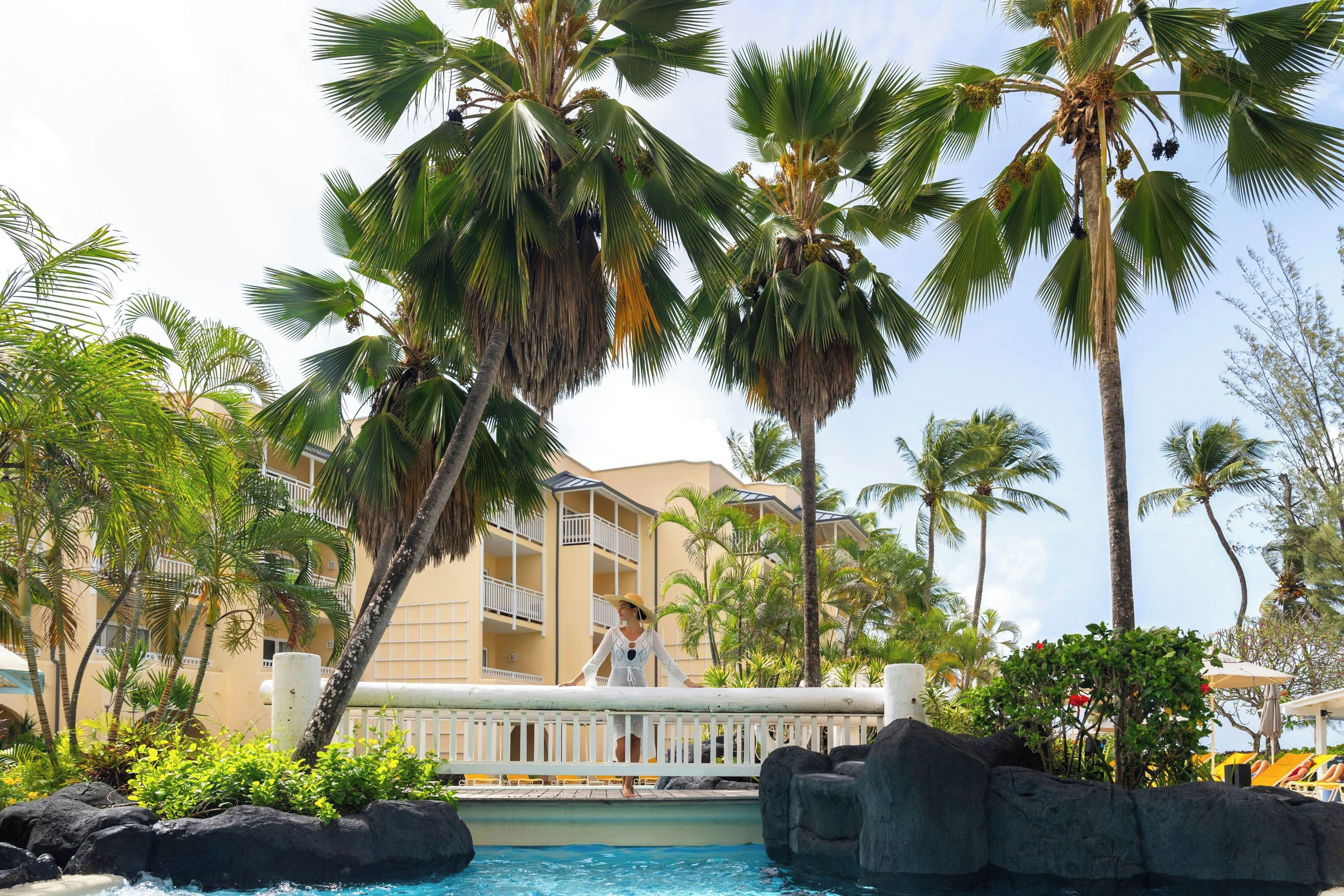Un área de piscina en un resort, con palmeras, una piscina y un edificio al fondo.