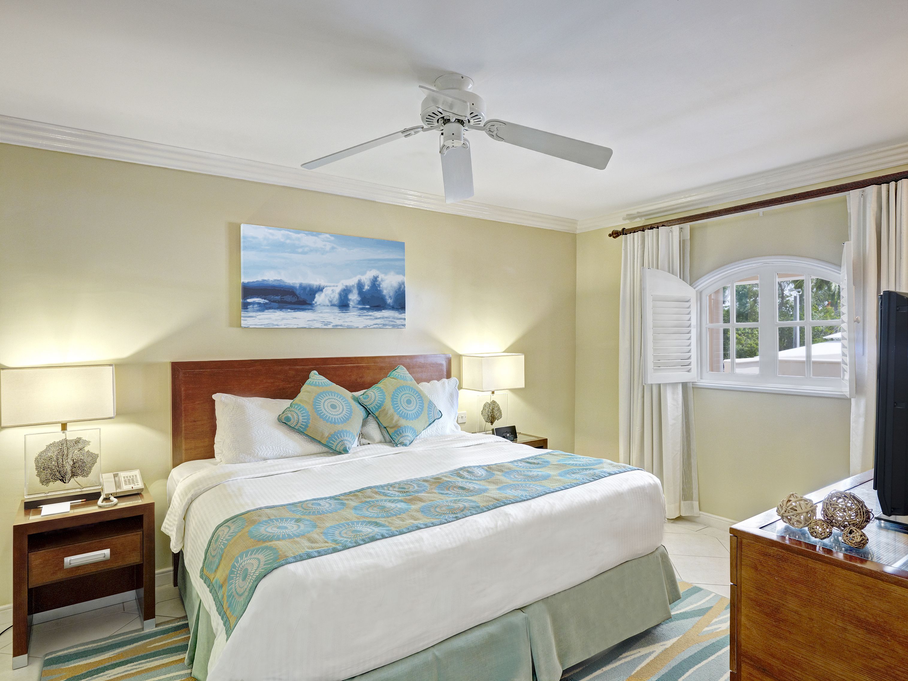 A hotel room with a bed, two bedside tables, a ceiling fan, a painting, and a window with curtains.