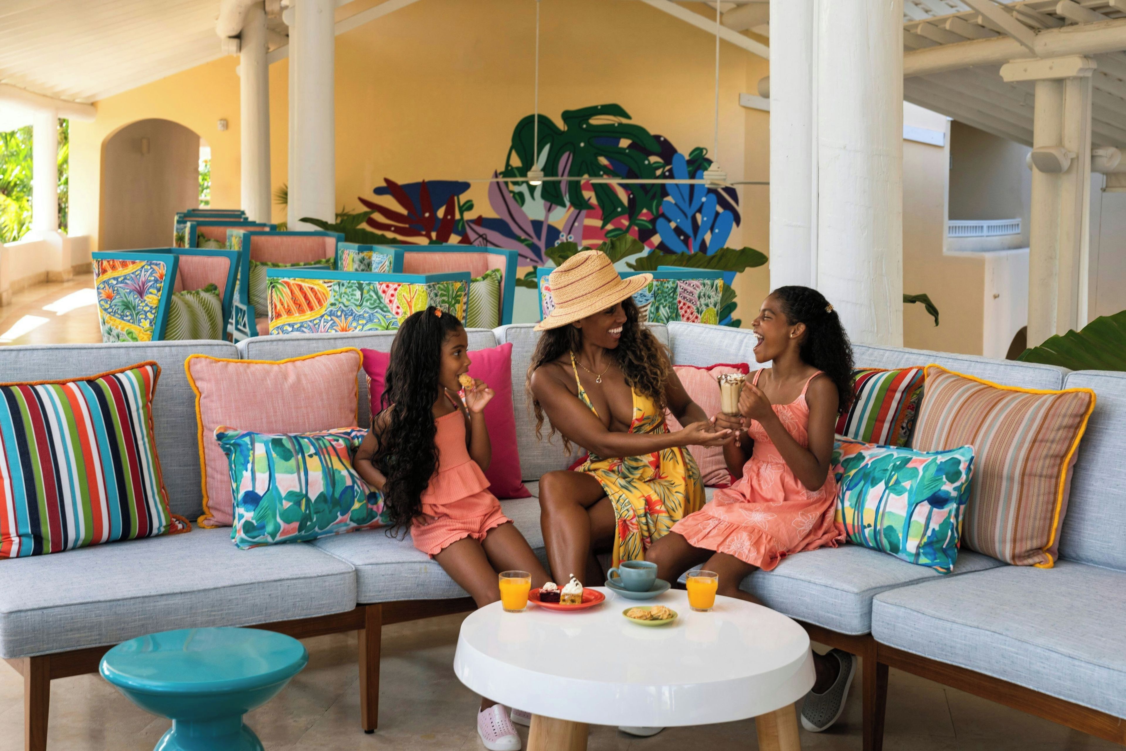 A seating area with a sofa, colorful cushions, a round table with food and drinks, and a tropical mural in the background.
