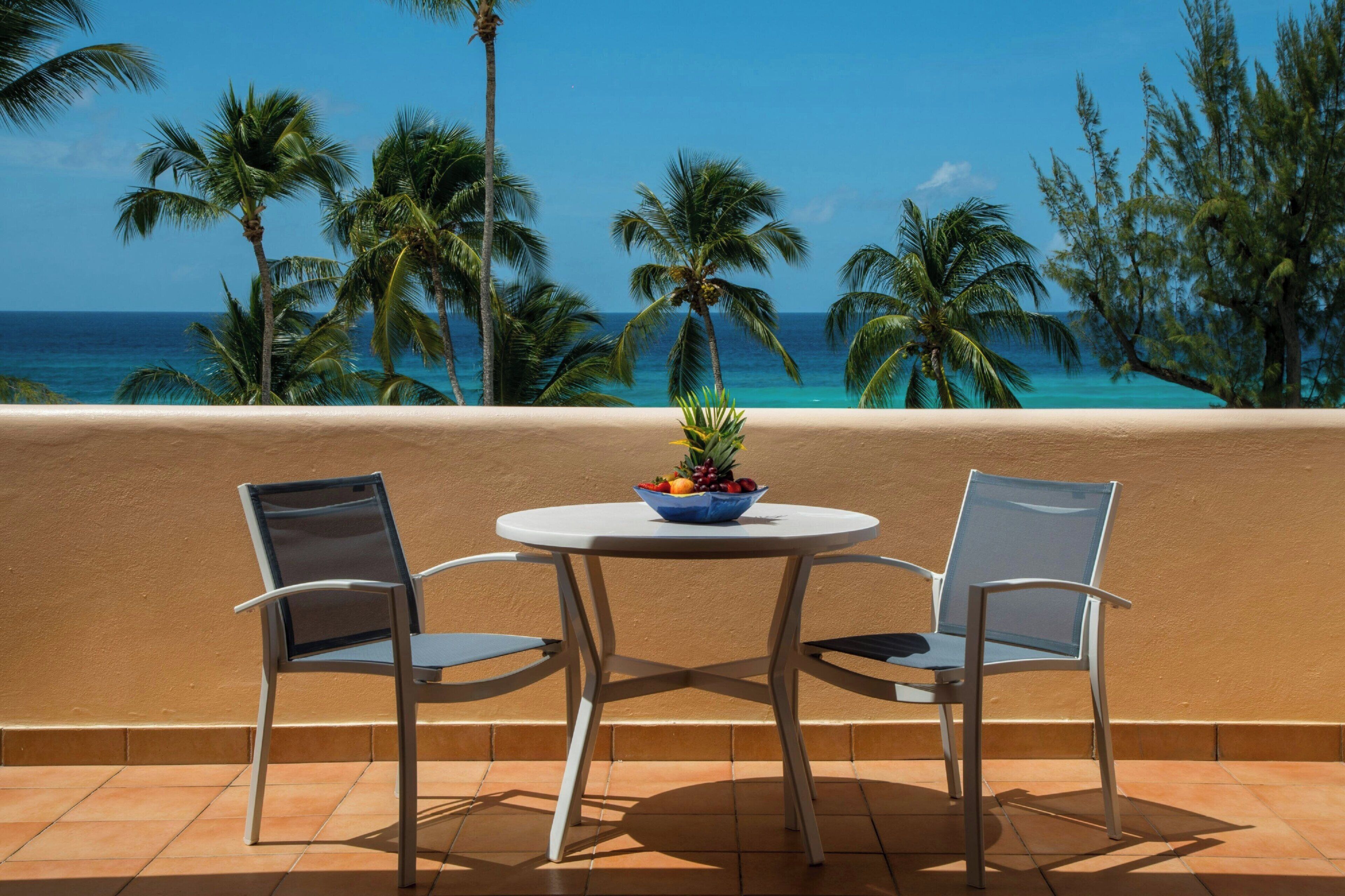 A balcony with two chairs and a table with a fruit bowl, overlooking a beach with palm trees and the ocean.