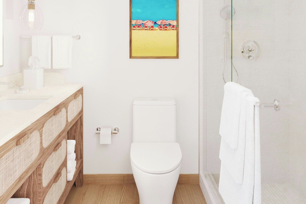 A modern bathroom with a wooden vanity, white fixtures, a glass shower enclosure, and a beach-themed painting on the wall.