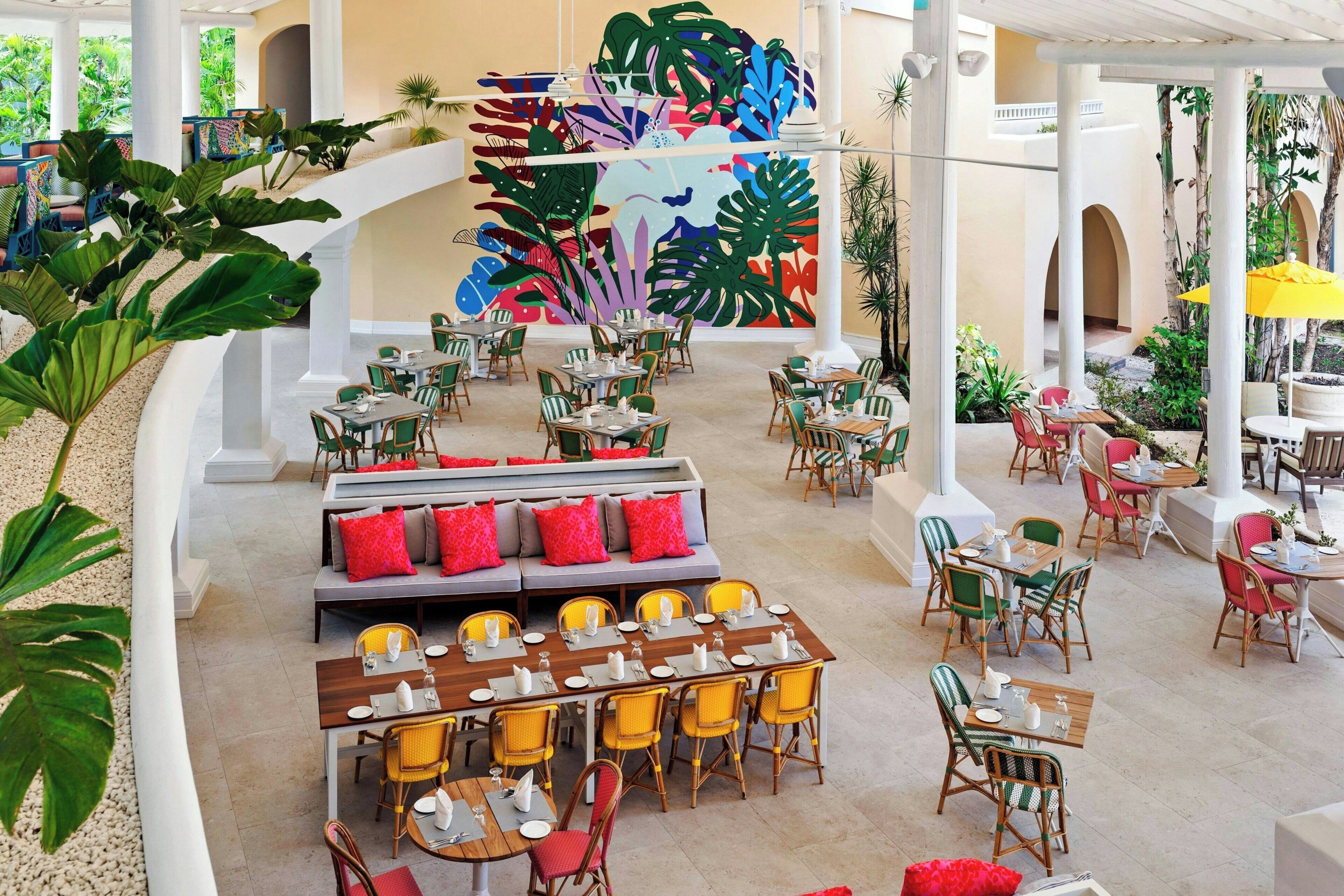 A spacious indoor area with multiple dining tables and chairs, a central seating area with a sofa, and a colorful mural on the wall.