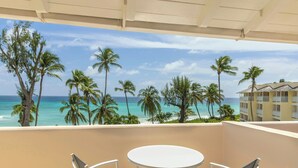 Un balcón con una mesa y sillas blancas, con vista a una playa con palmeras y un cielo azul despejado.