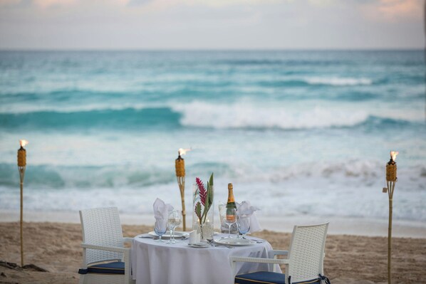 A table set for two on a beach with ocean views, candles, and a bottle of champagne.