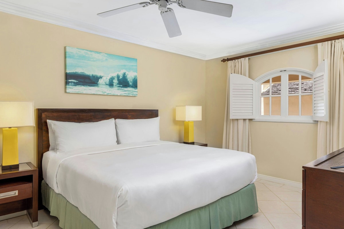 Habitación de hotel con una cama, dos lámparas de noche, un ventilador de techo, una ventana con cortinas y un cuadro en la pared.