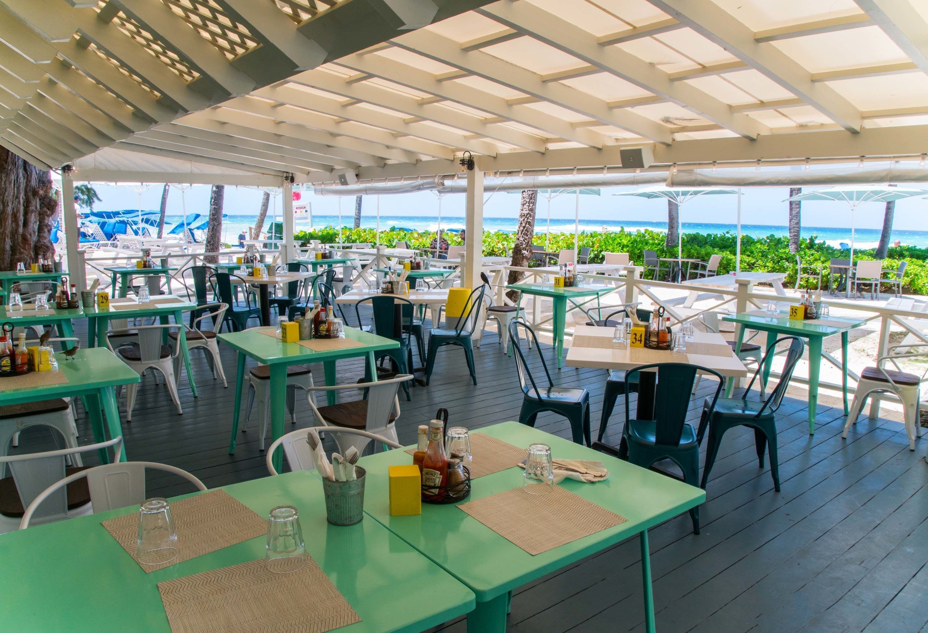 An outdoor dining area with tables and chairs, a view of the beach, and a covered section with a white ceiling.
