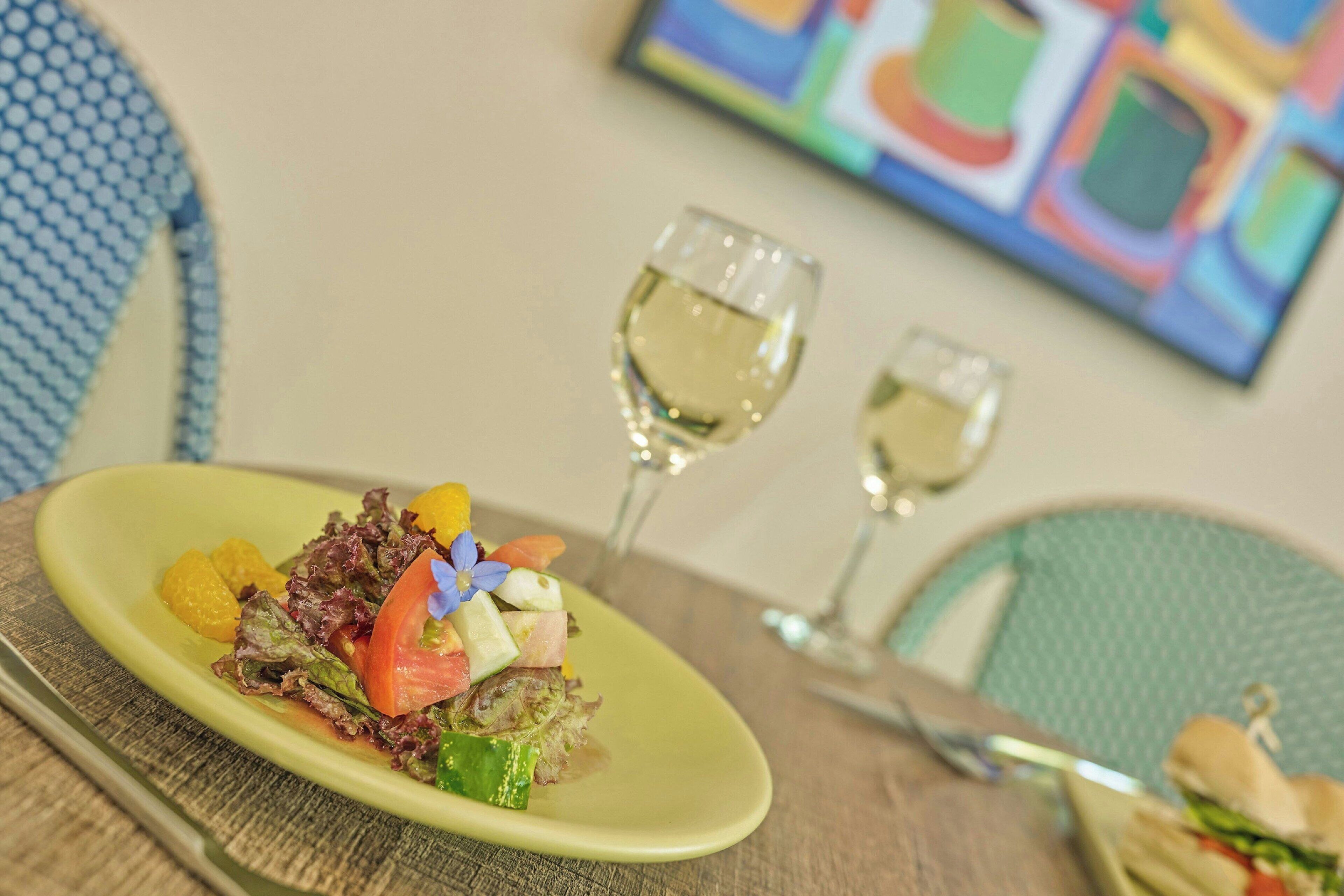 A dining table with a plate of food, two glasses of white wine, and a colorful painting in the background.