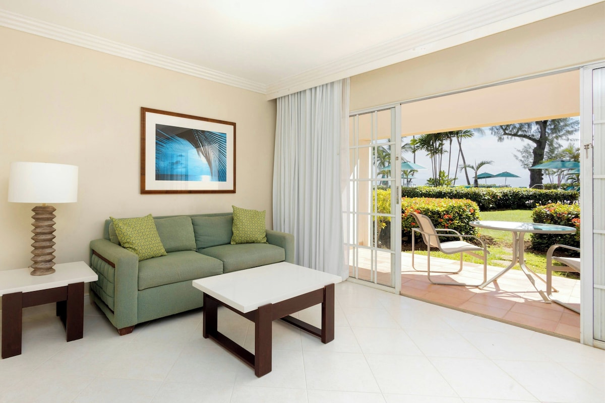 Una habitación moderna con un sofá verde, una mesa blanca y una puerta de cristal que da a un patio exterior con sillas y una mesa.