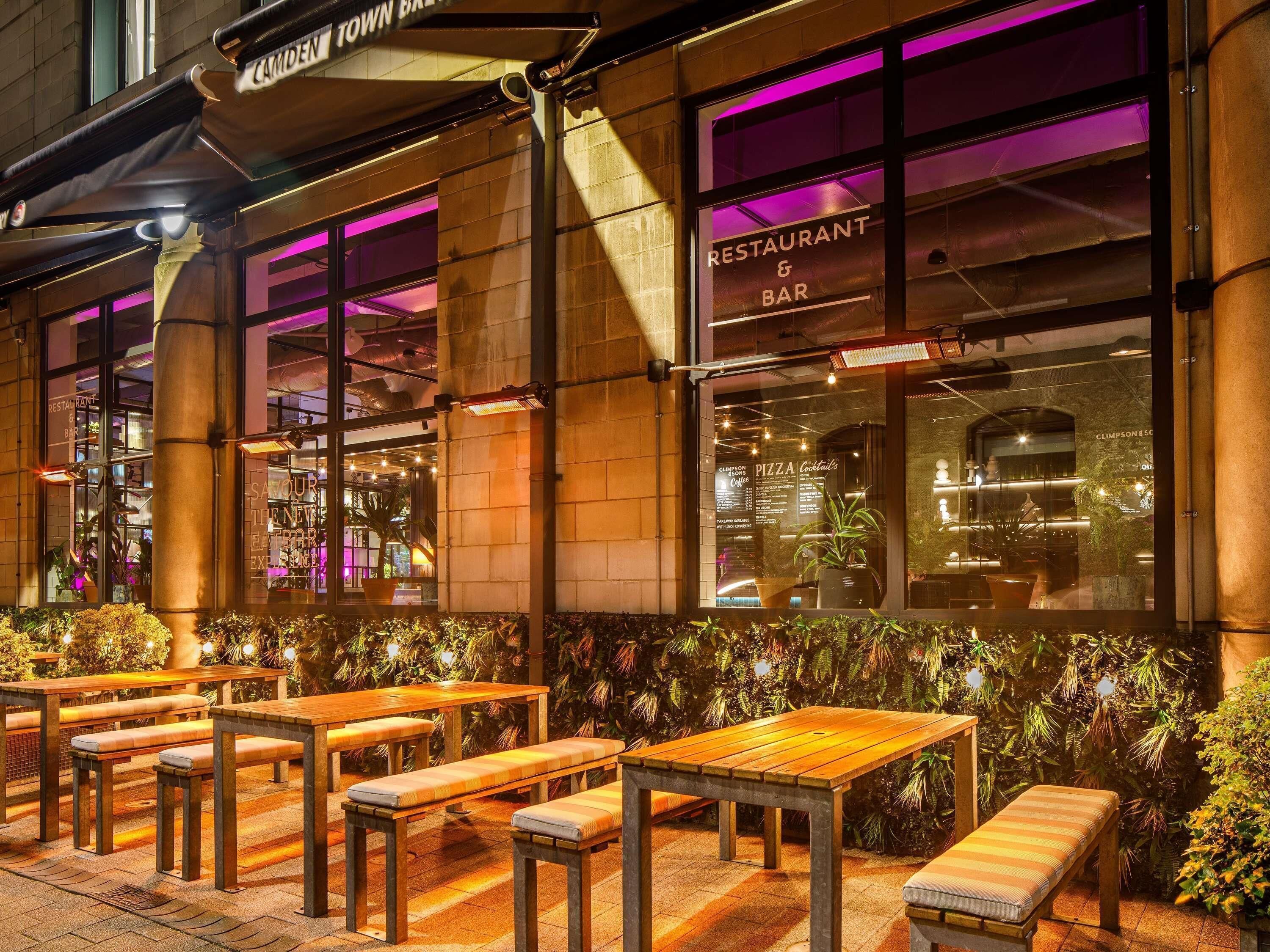 An outdoor seating area with wooden tables and benches, a sign for a restaurant and bar, and a view of the interior through the windows.
