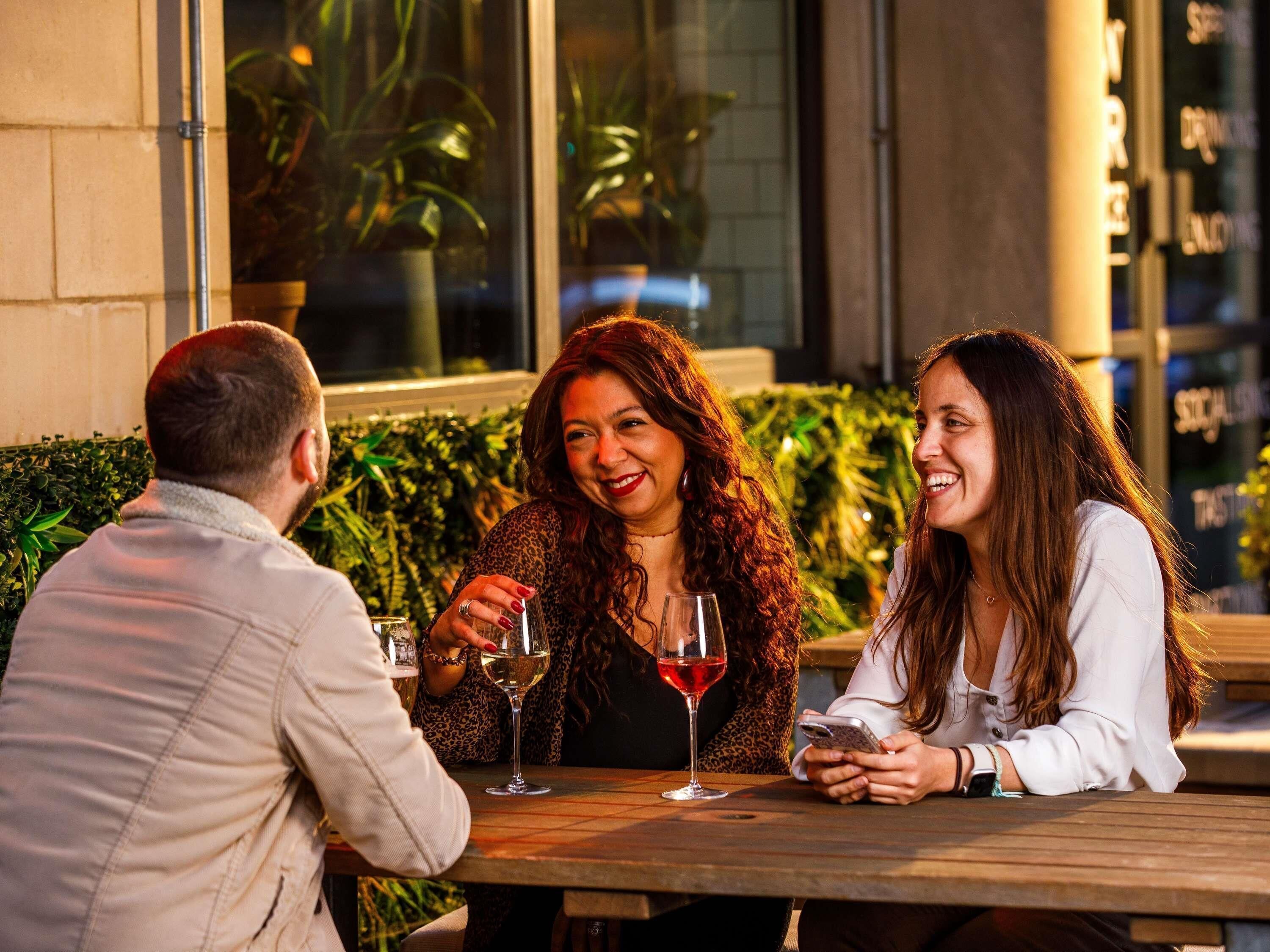 Three people seated at an outdoor table with drinks, in front of a building with large windows and greenery.