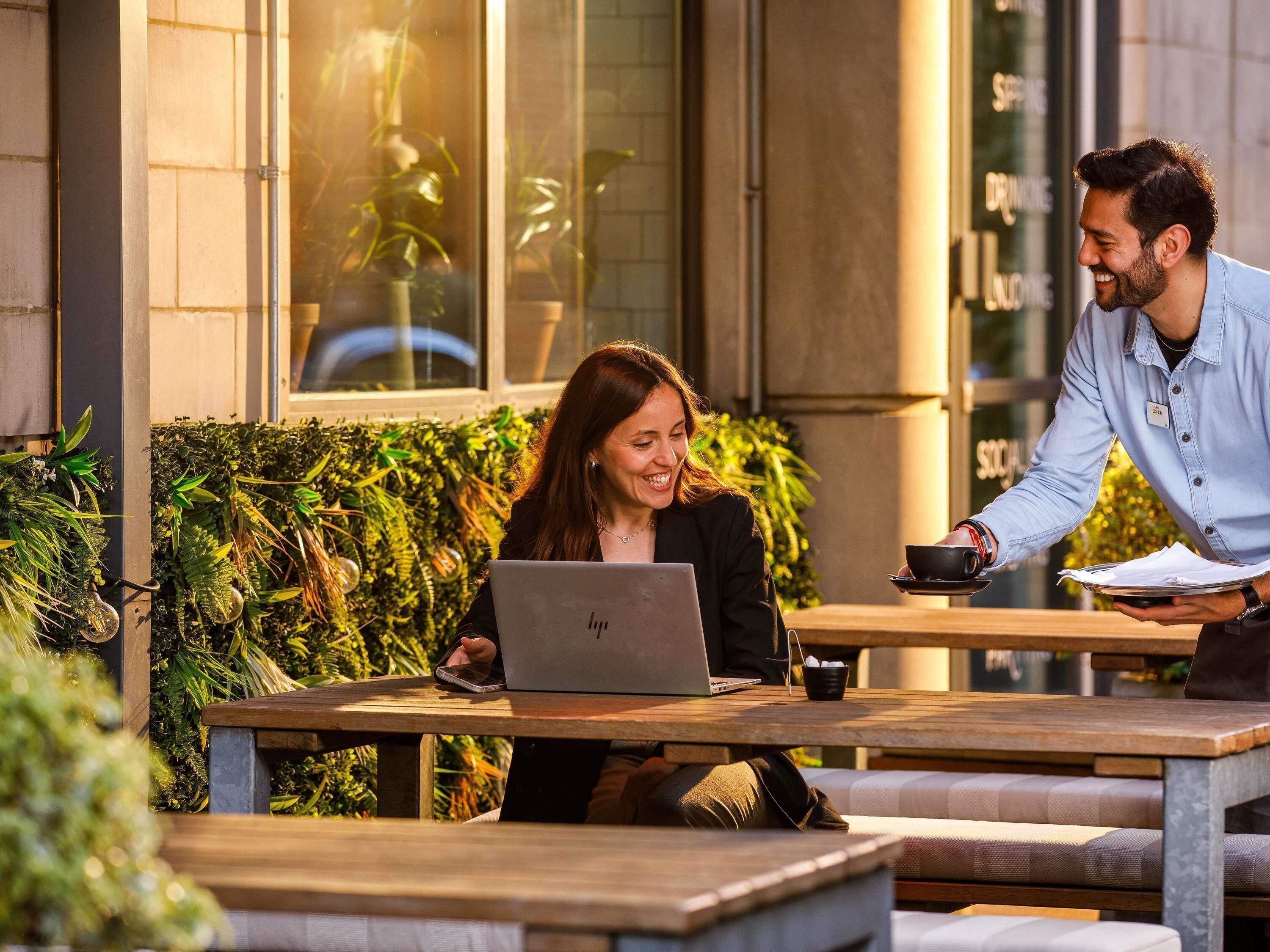 An outdoor seating area with tables and benches, a laptop on one table, and a person holding a cup and papers.