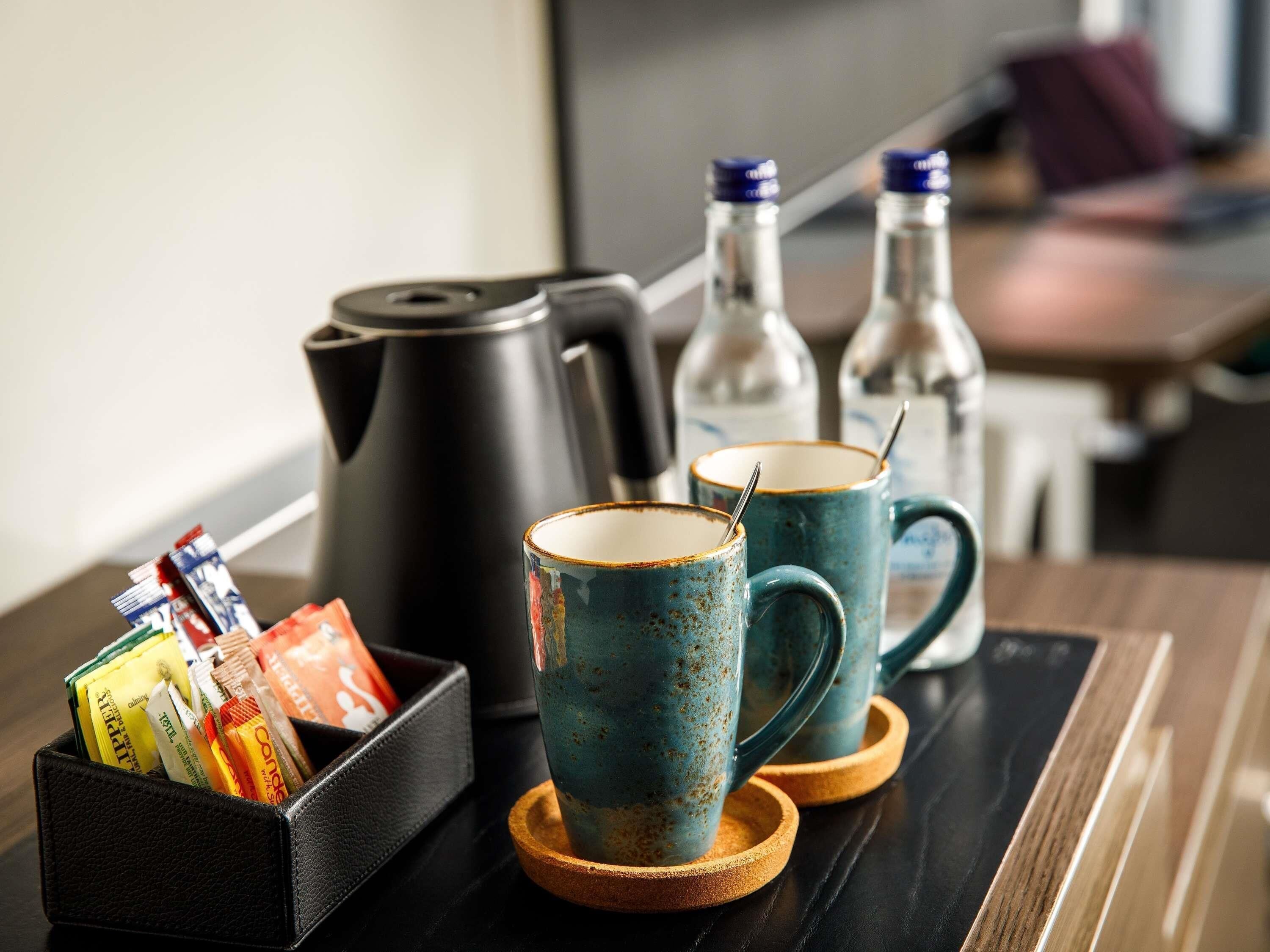 A coffee station with a black coffee pot, two teal mugs with spoons, and a box of assorted coffee pods.