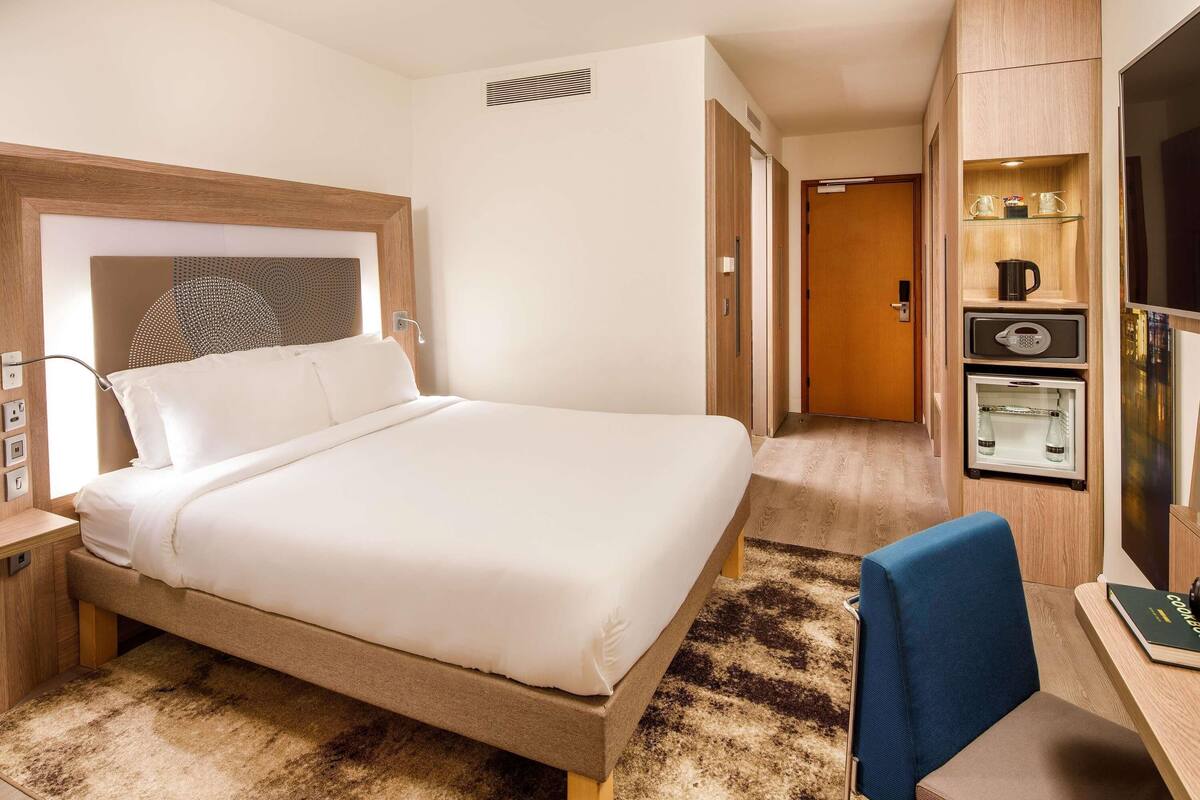 A hotel room with a bed, a desk, a chair, a minibar, and a television.