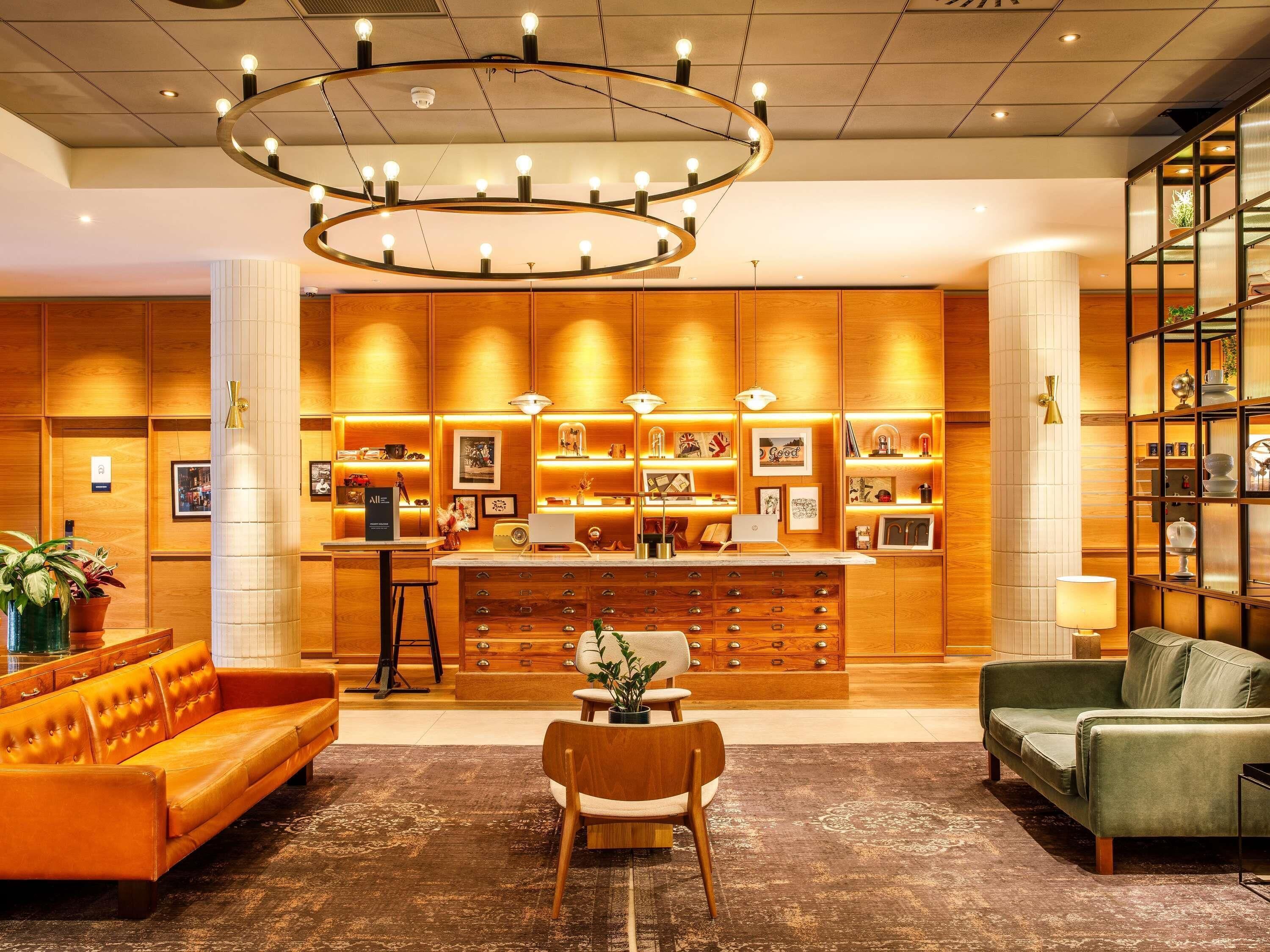 A hotel lobby with a central reception desk, wooden paneling, a large chandelier, and seating areas.