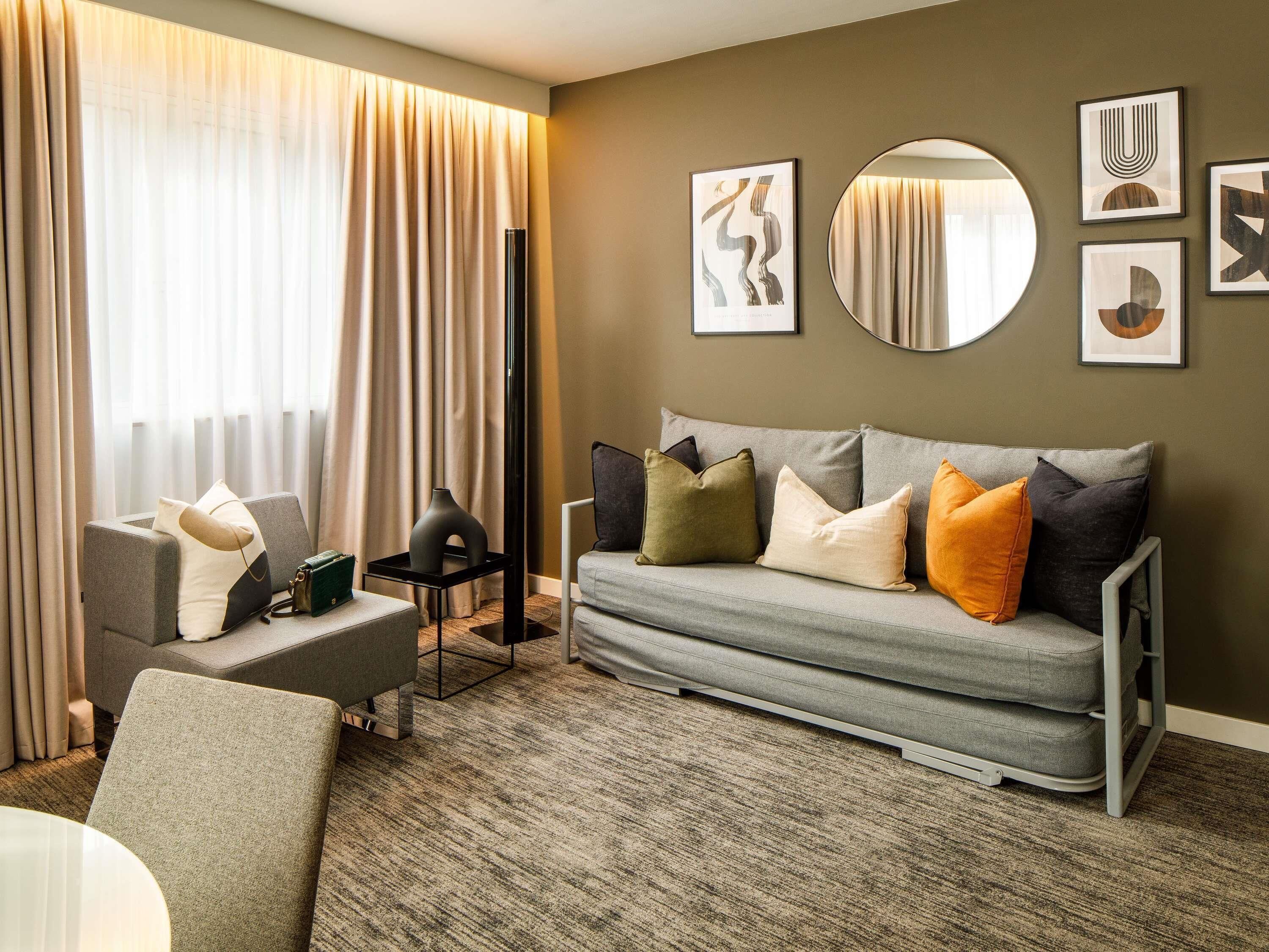 Quarto de hotel moderno com um sofá cinzento, um espelho redondo e arte abstrata na parede.