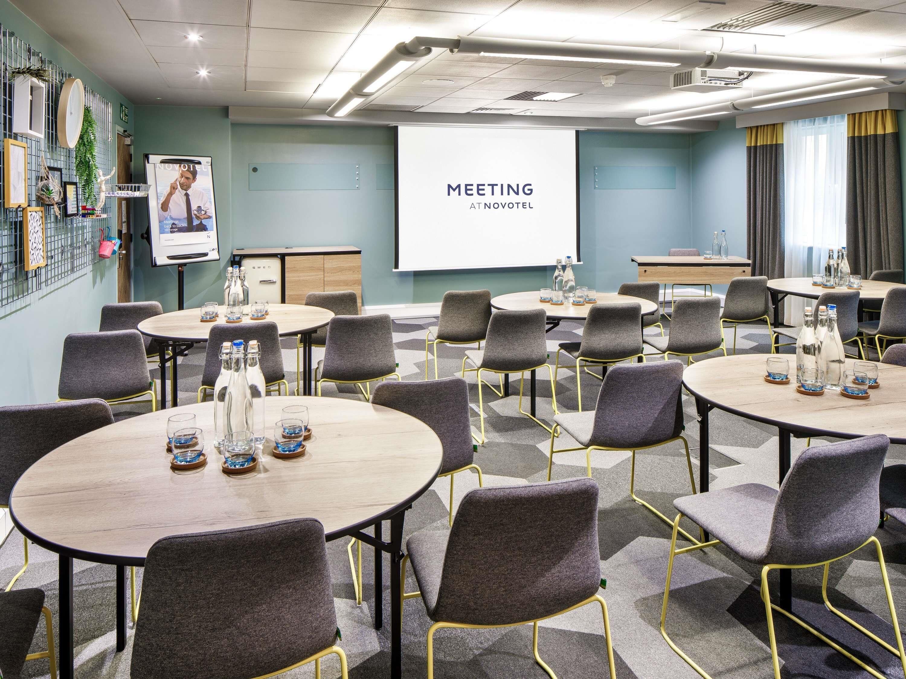 A meeting room with round tables, grey chairs, and a projector screen displaying 'MEETING AT NOVOTEL'.