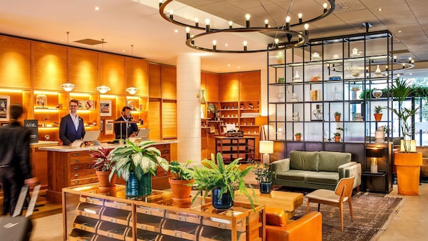 A hotel lobby with a reception desk, a circular chandelier, a green sofa, and a bookshelf with potted plants.