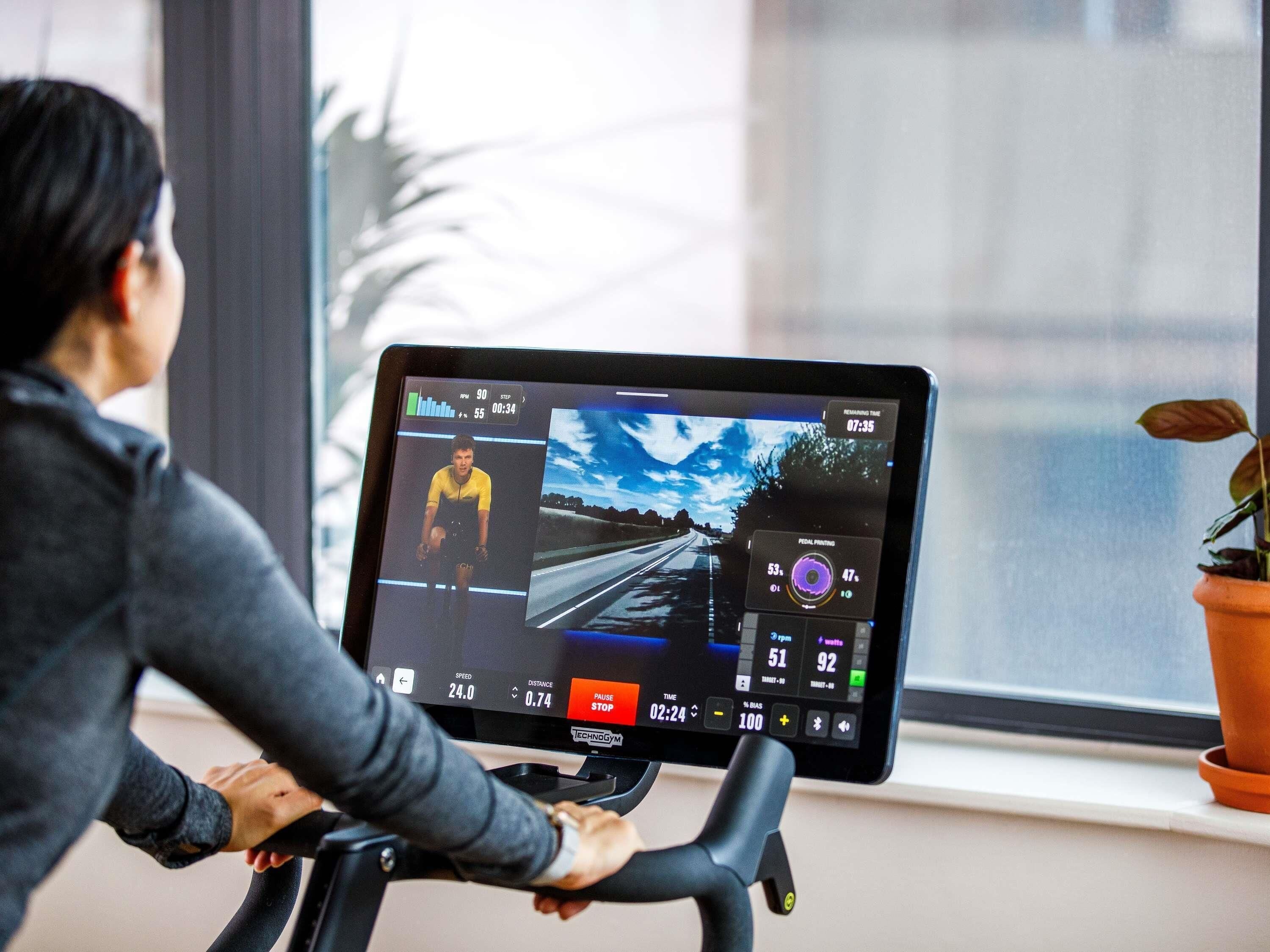 Una persona usando una bicicleta estática con una pantalla digital, una ventana con una planta y una vista de carretera en la pantalla.