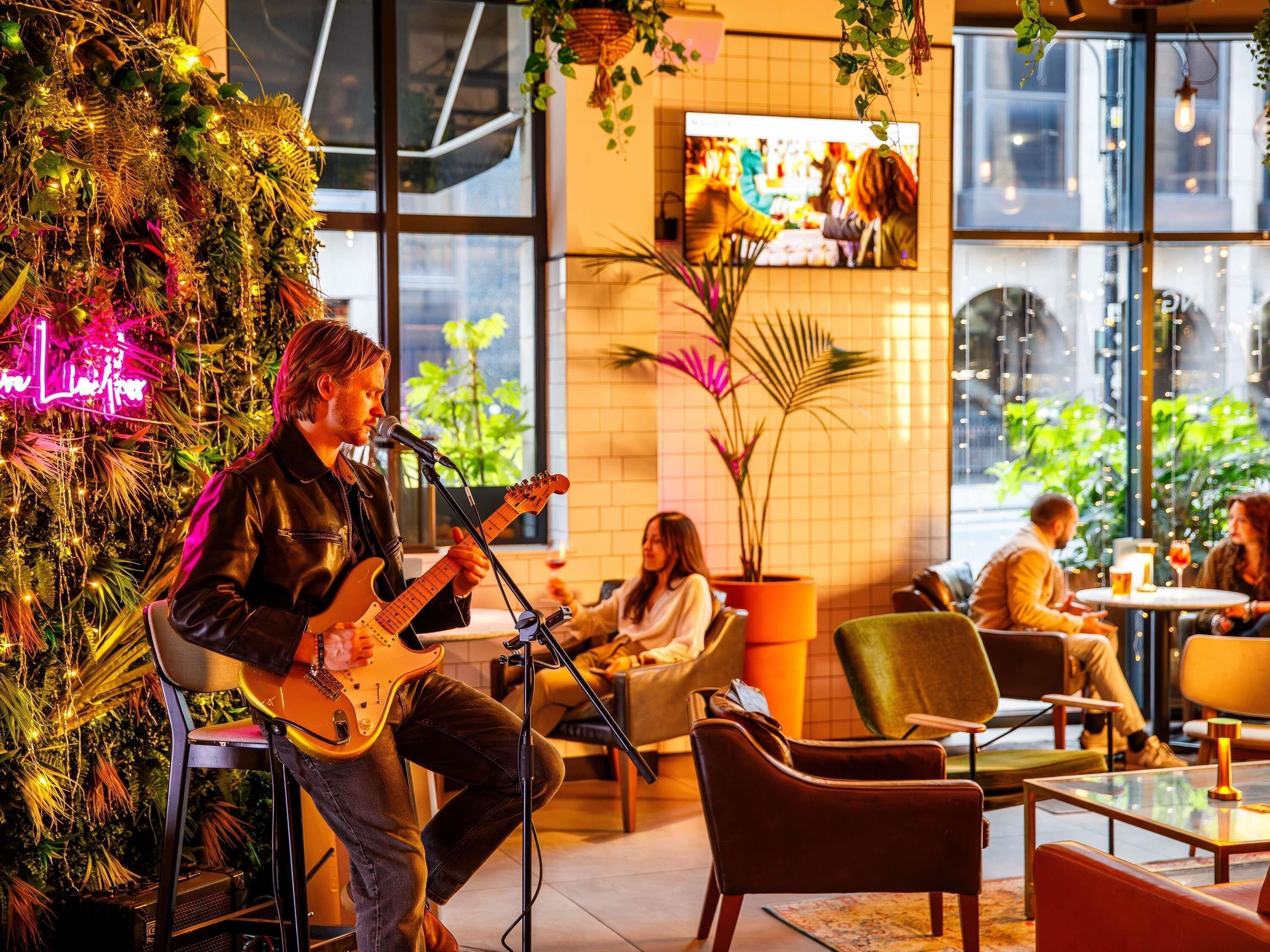 Un musicista che si esibisce in un ambiente interno accogliente, con una pianta grande e un neon sullo sfondo.