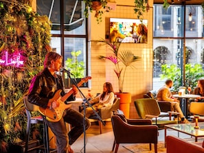 A musician performing in a cozy indoor setting with a large plant and a neon sign in the background.